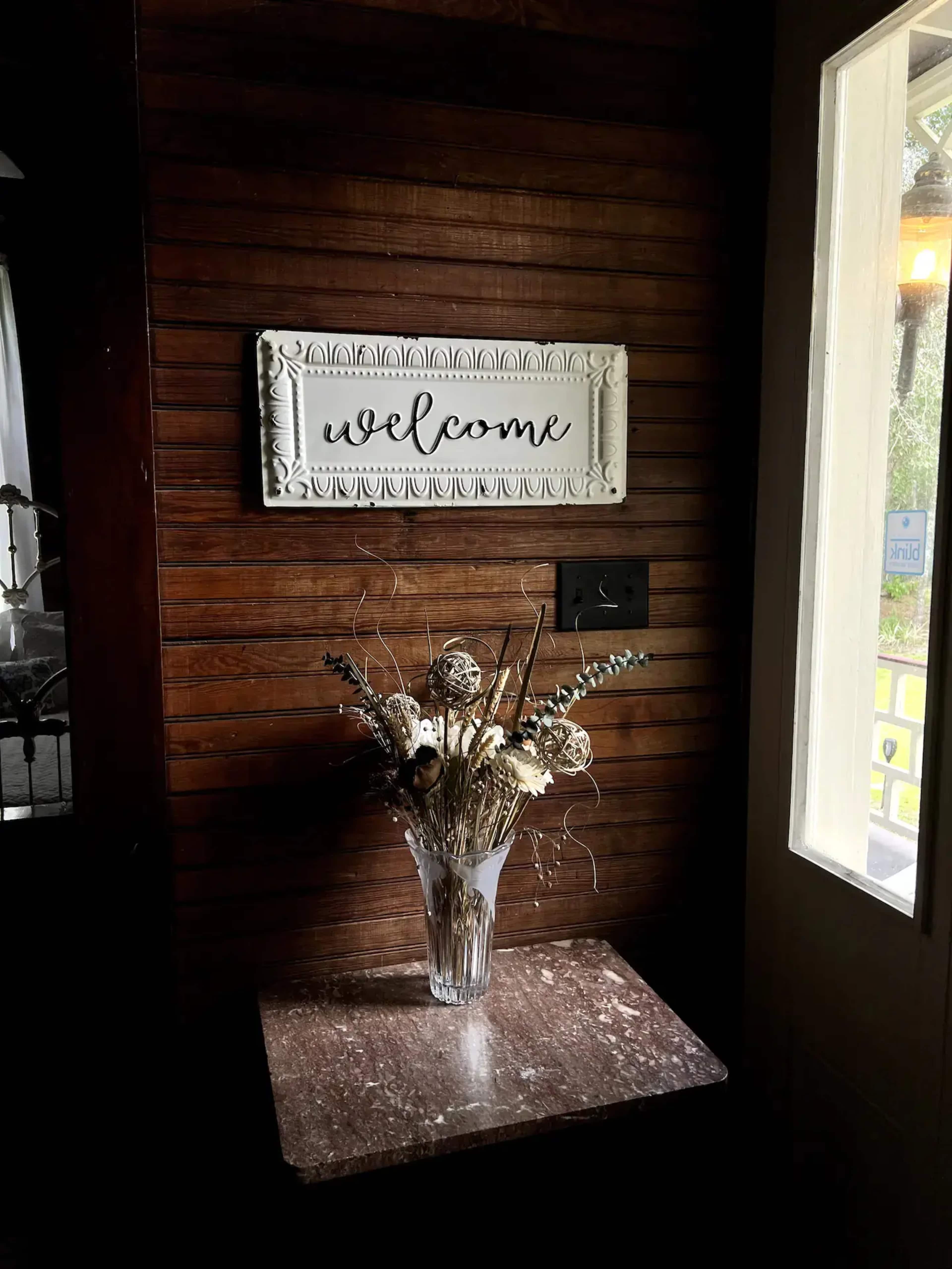 A "welcome" sign is mounted on a wooden wall above a table holding a vase with dried flowers.