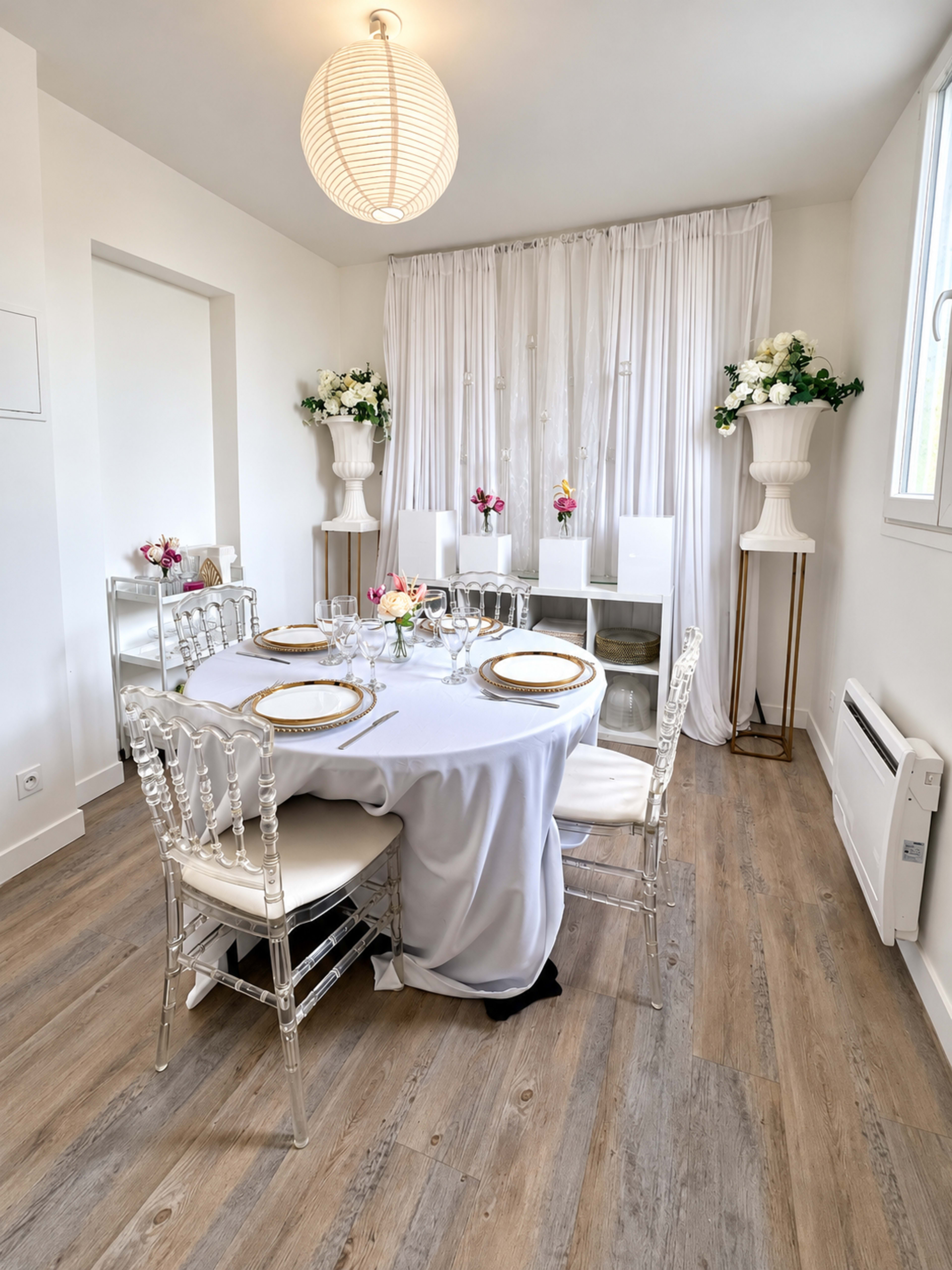 A dining area features a round table set with elegant dinnerware, surrounded by transparent chairs, and adorned with floral arrangements and a soft white curtain backdrop.