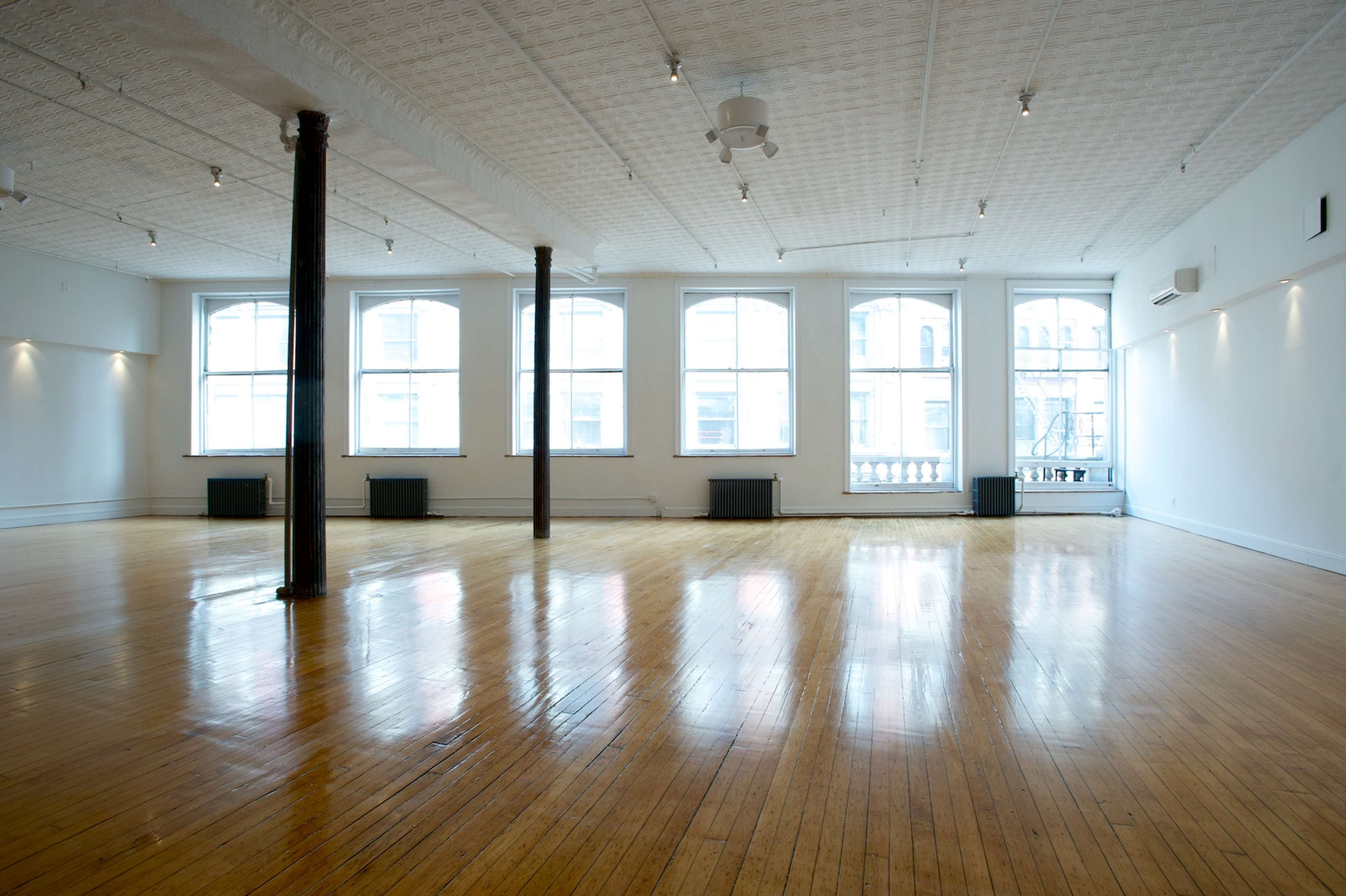 A spacious, empty room with polished wooden floors and large windows allowing natural light to fill the space.
