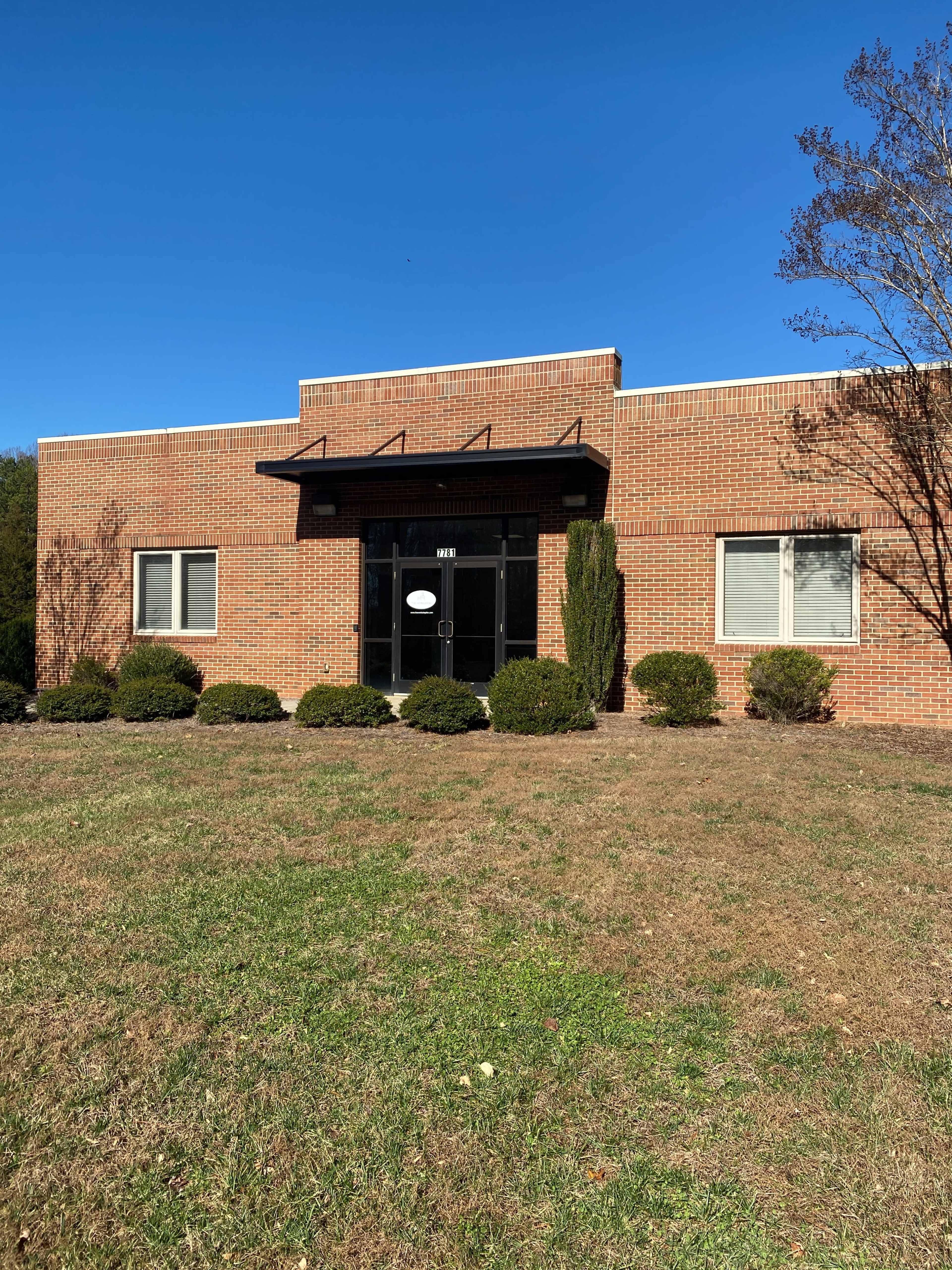 A brick building with a black entrance door and two large windows on either side, surrounded by neatly trimmed bushes and a clear blue sky.