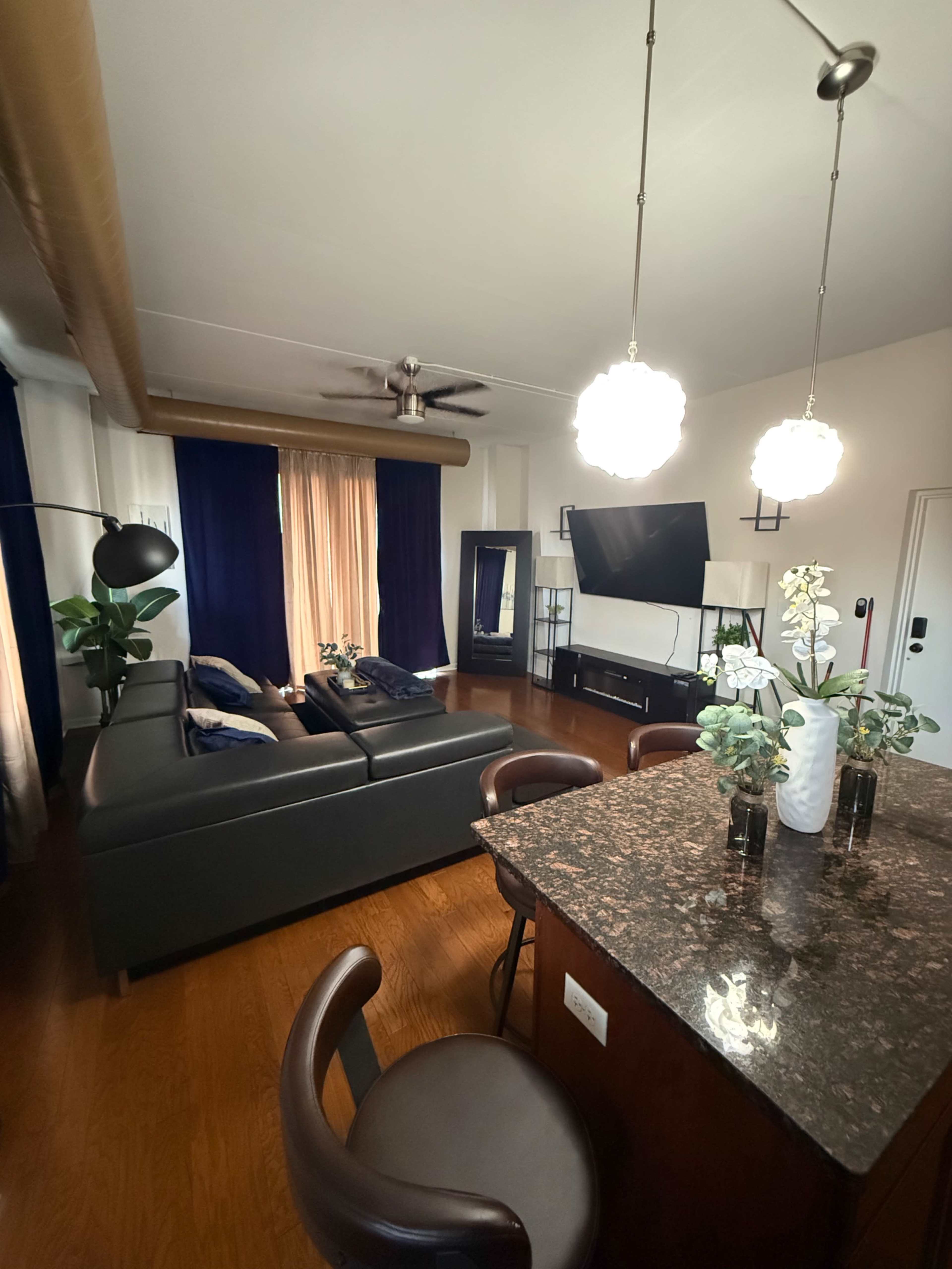 The image shows a modern living room with a dark sectional sofa, a television mounted on the wall, and a dining area featuring barstool seating around a granite island.