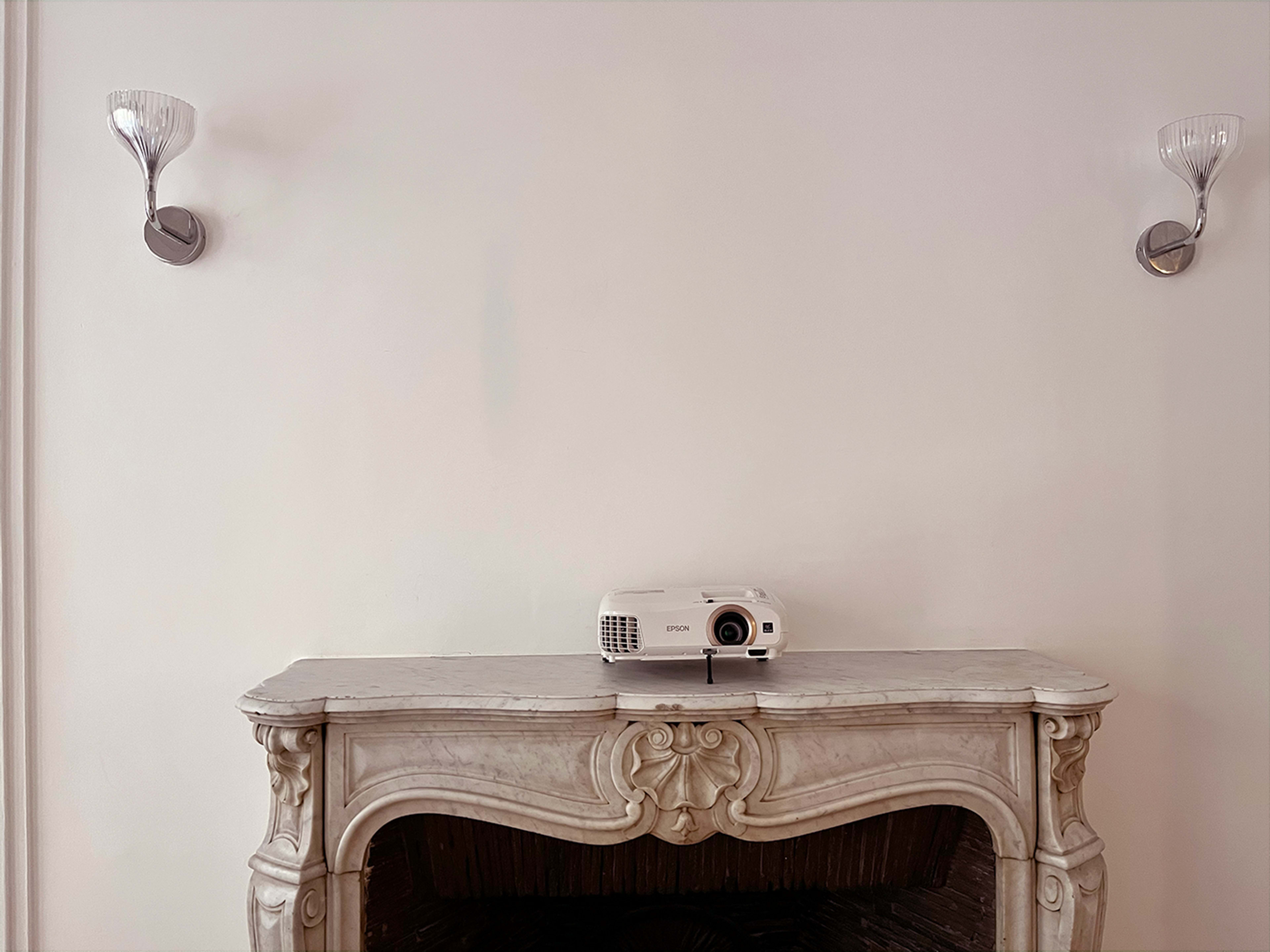 A white projector is placed on a marble fireplace beneath two wall sconces.
