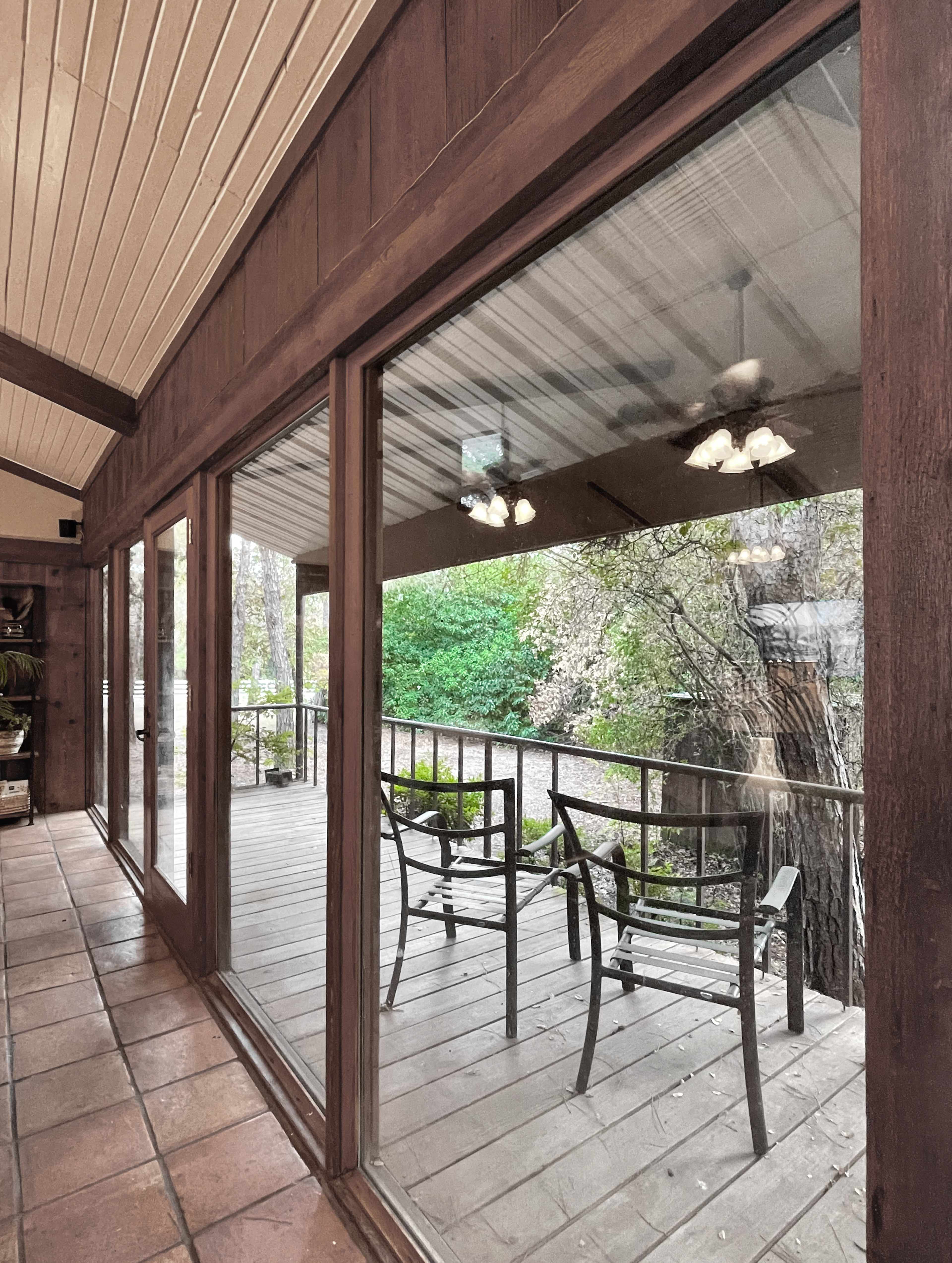 A view through large glass doors onto a wooden deck with two chairs and greenery in the background.