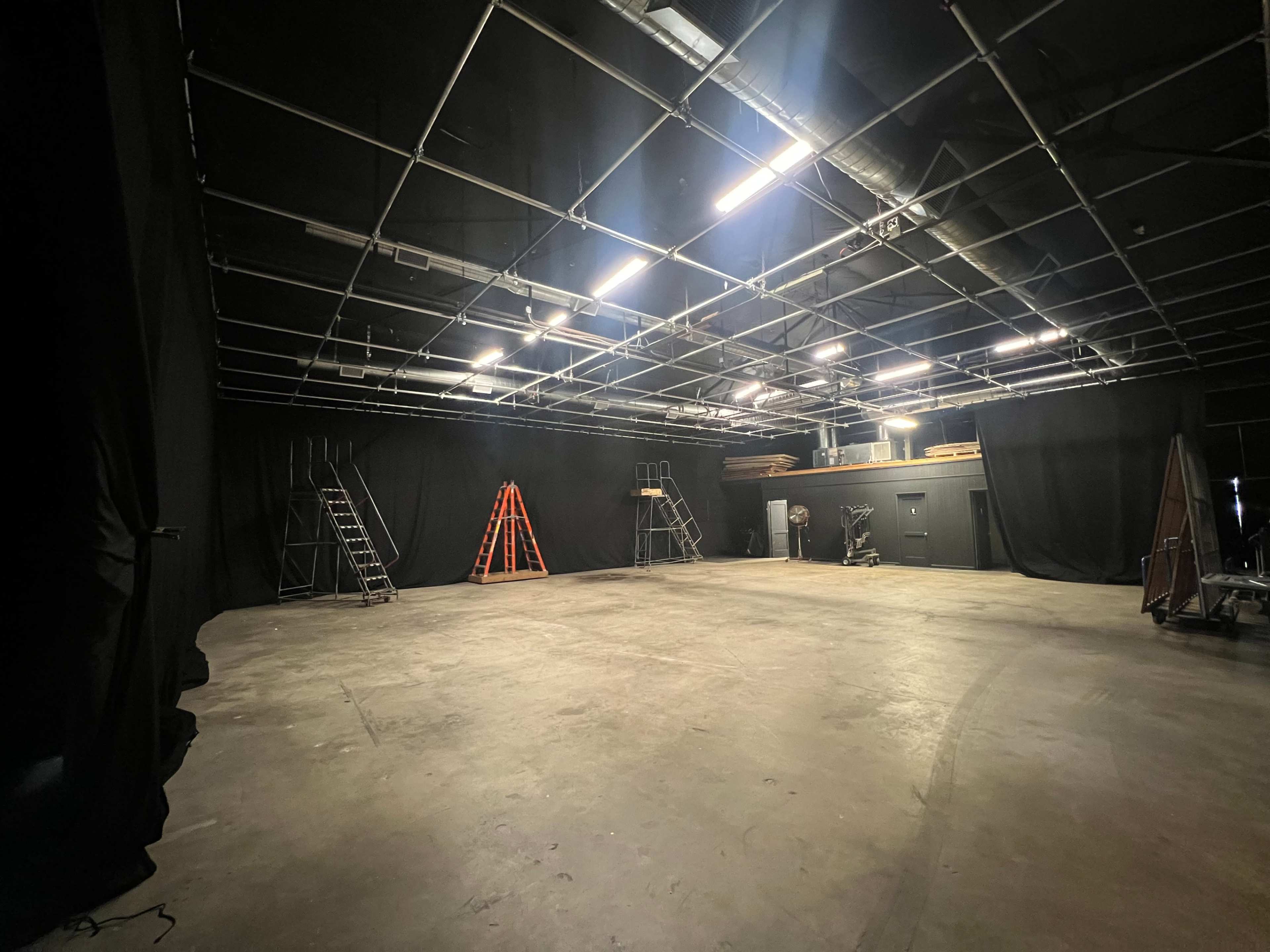 An empty studio space features black curtains lining the walls, several step ladders, and an assortment of equipment arranged in the back.