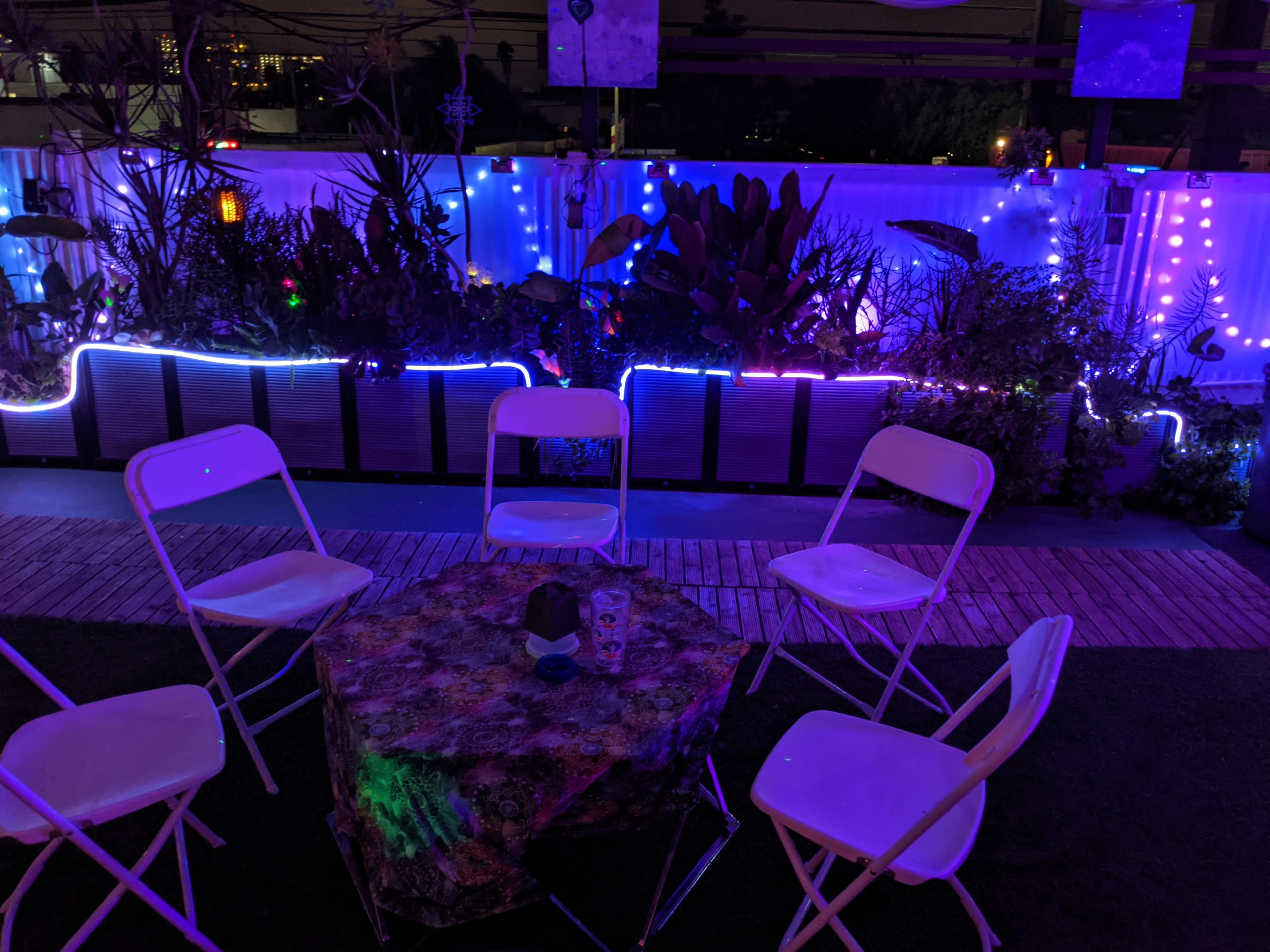 The scene depicts a rooftop area with four white chairs arranged around a table, surrounded by plants and illuminated with purple and blue lights.