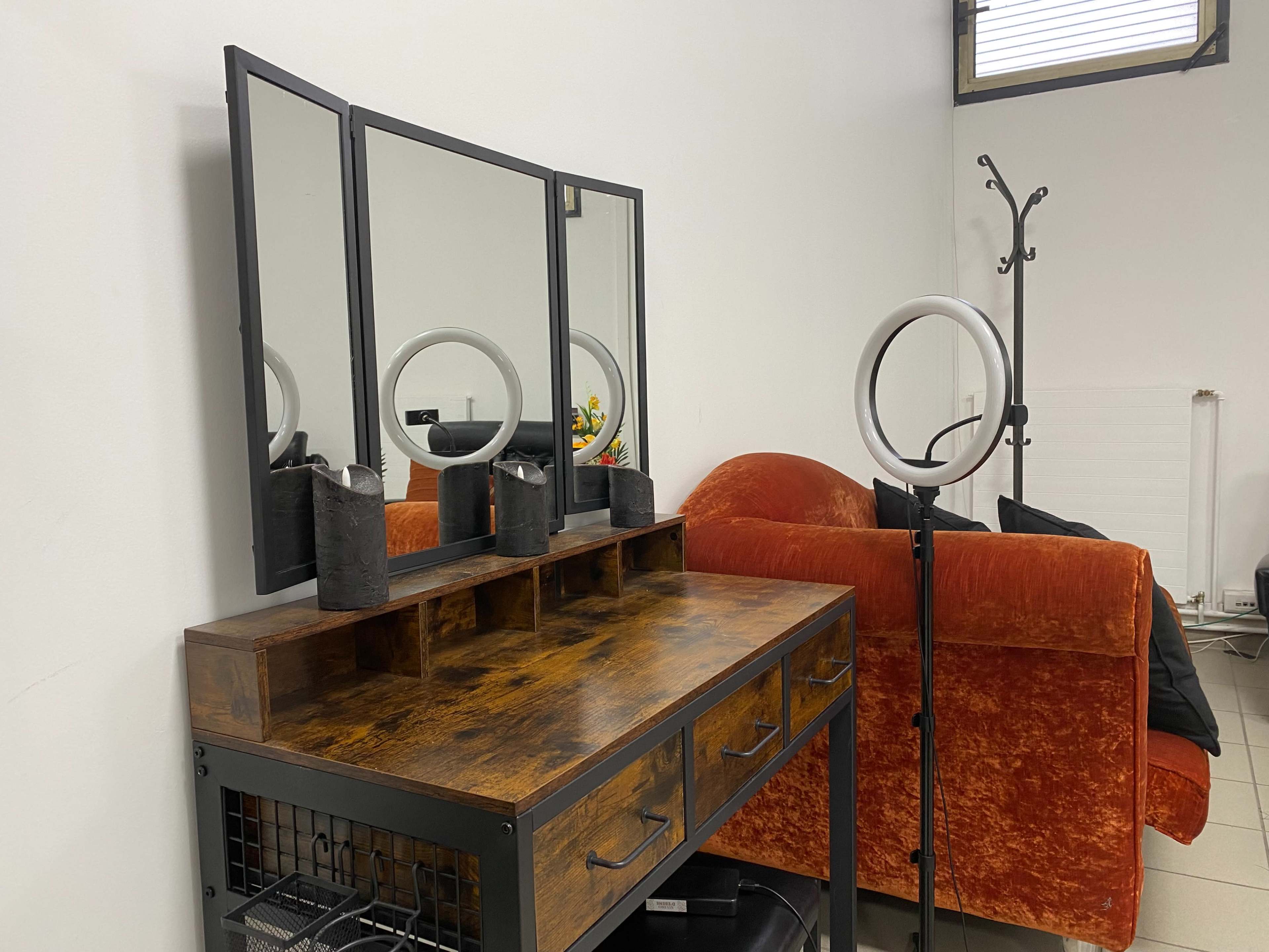 A wooden vanity with a three-panel mirror and decorative items is positioned next to an orange sofa in a minimalist room.