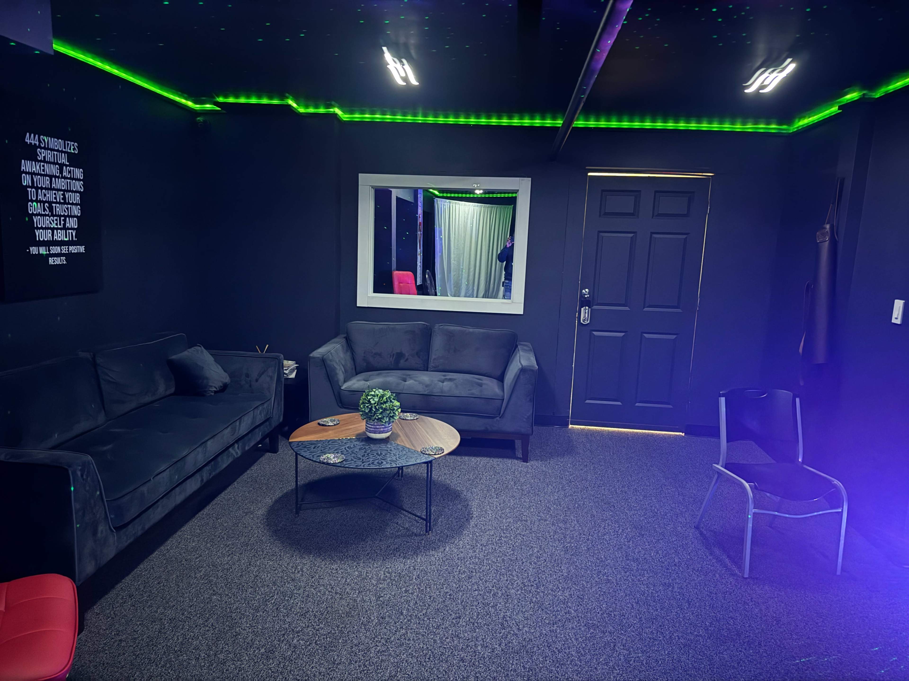 The image shows a dimly lit lounge area with dark walls, a mirror, two gray sofas, a coffee table, and a door.