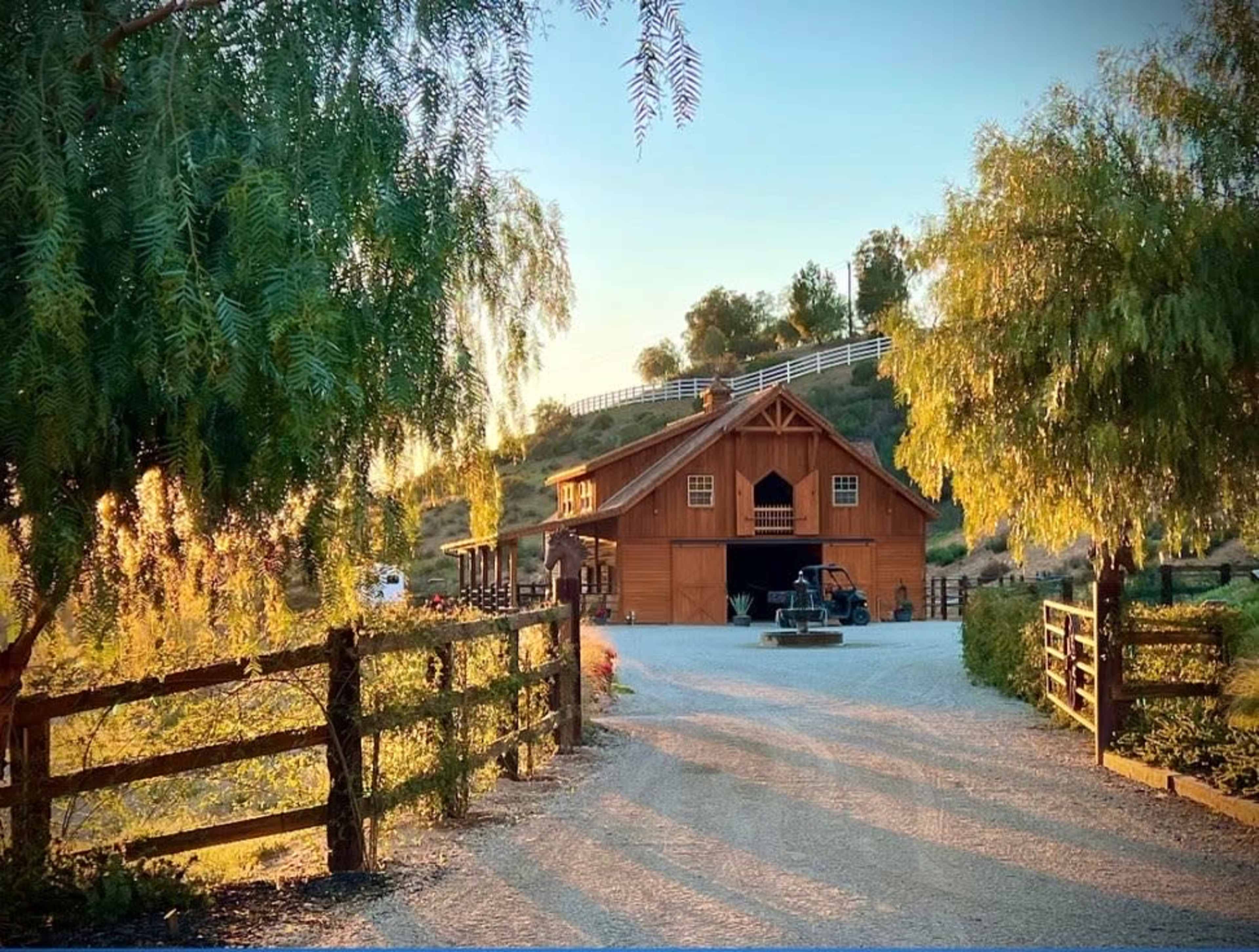 A wooden barn is nestled among trees at the end of a gravel driveway, leading to a hilly landscape.