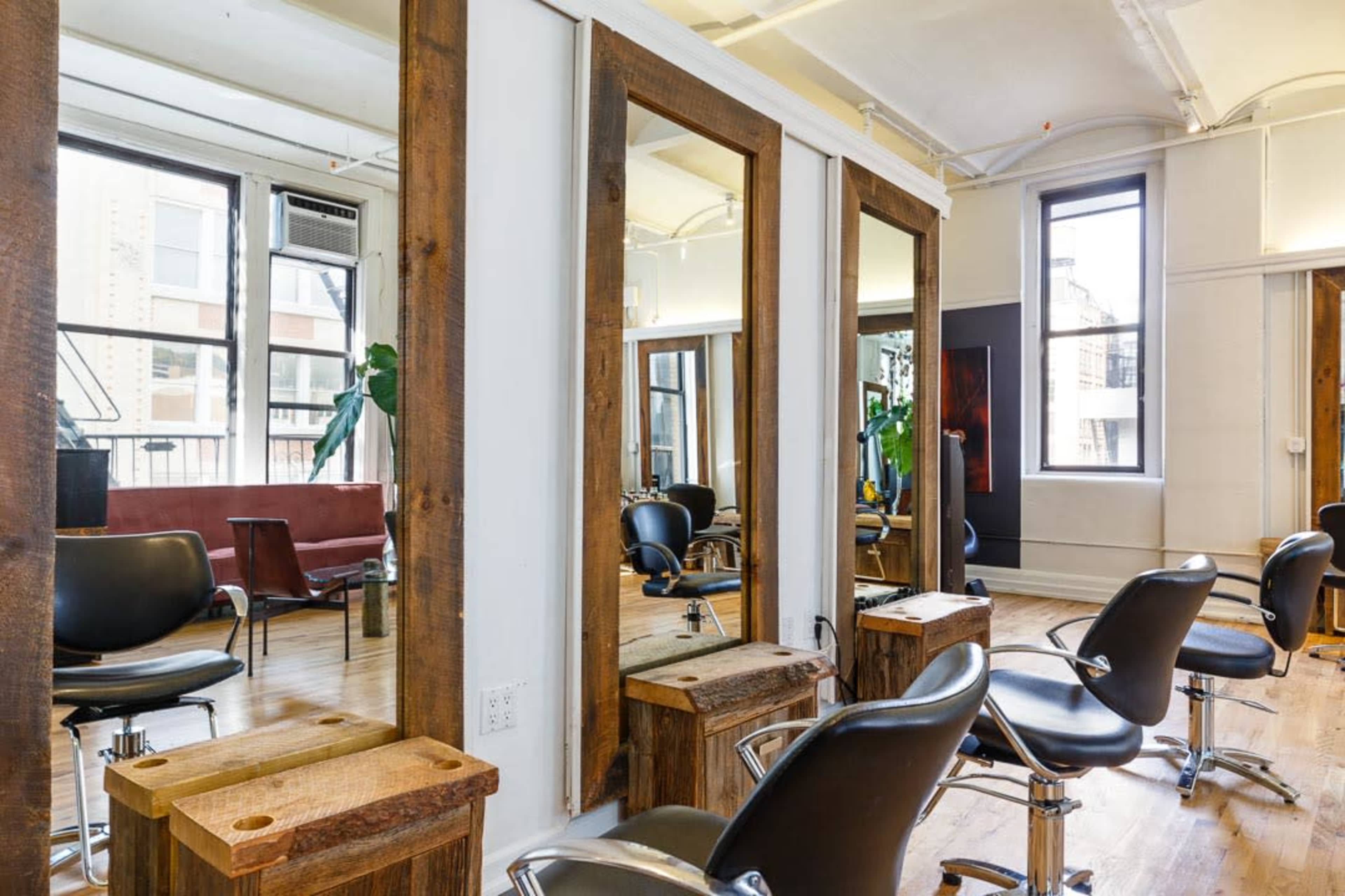 The image shows a modern hair salon with wooden-framed mirrors, black salon chairs, and a waiting area with a red sofa.