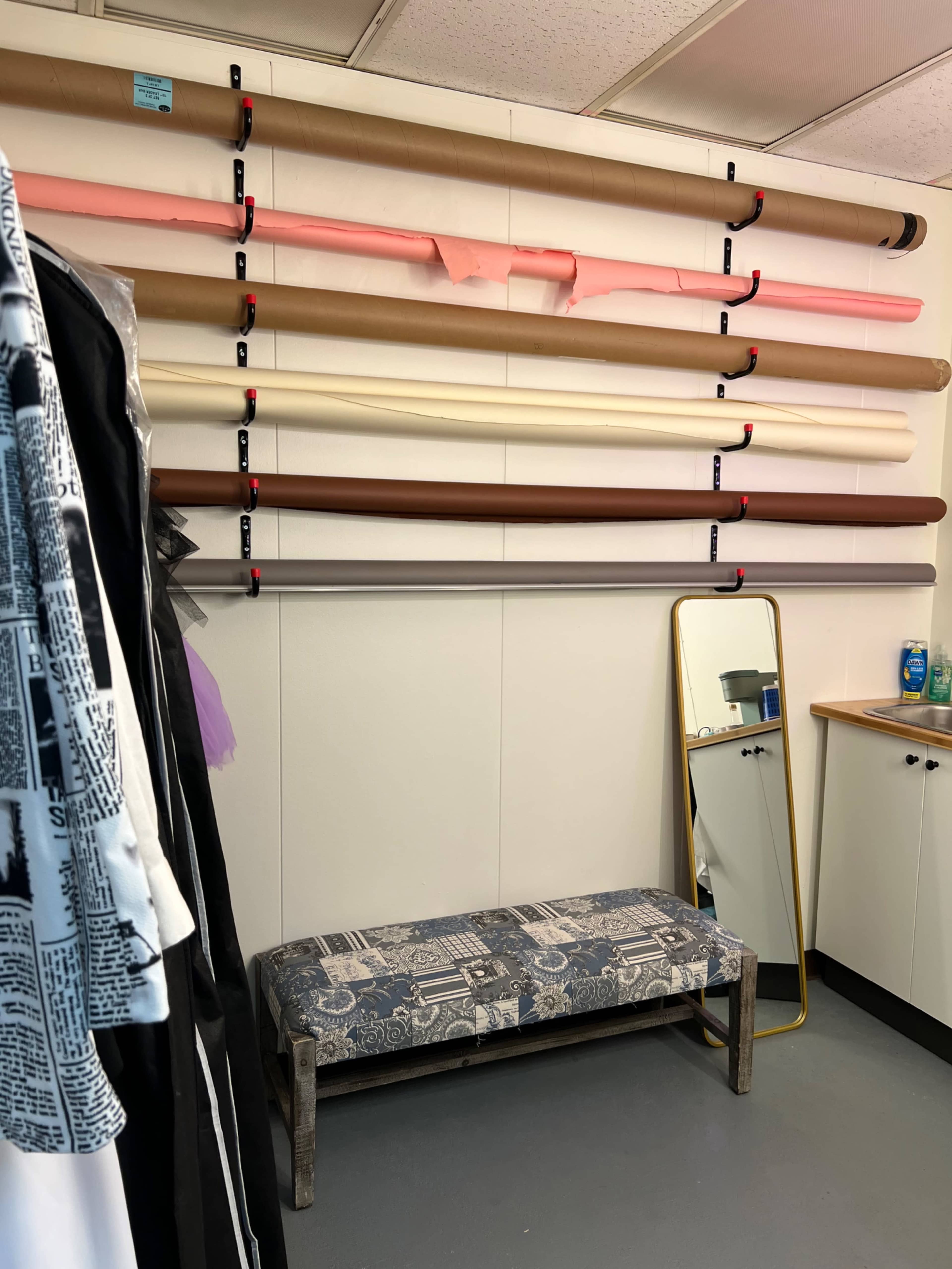 The image shows a small studio space with rolls of various colored paper organized on a wall above a wooden bench and a mirror.