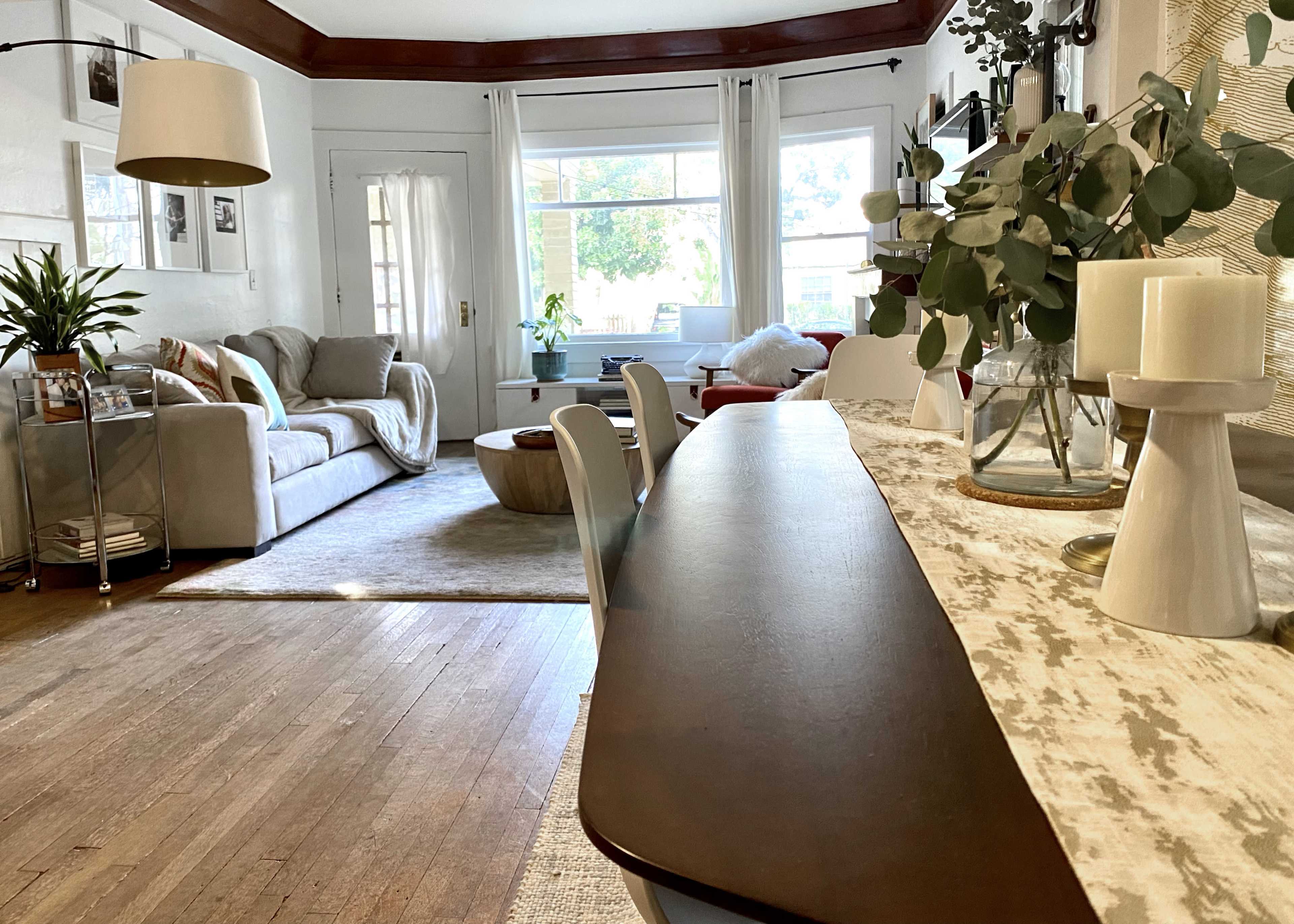 The image shows a cozy living space featuring a dining table with a table runner, a light-colored sofa, and large windows allowing natural light to enter the room.