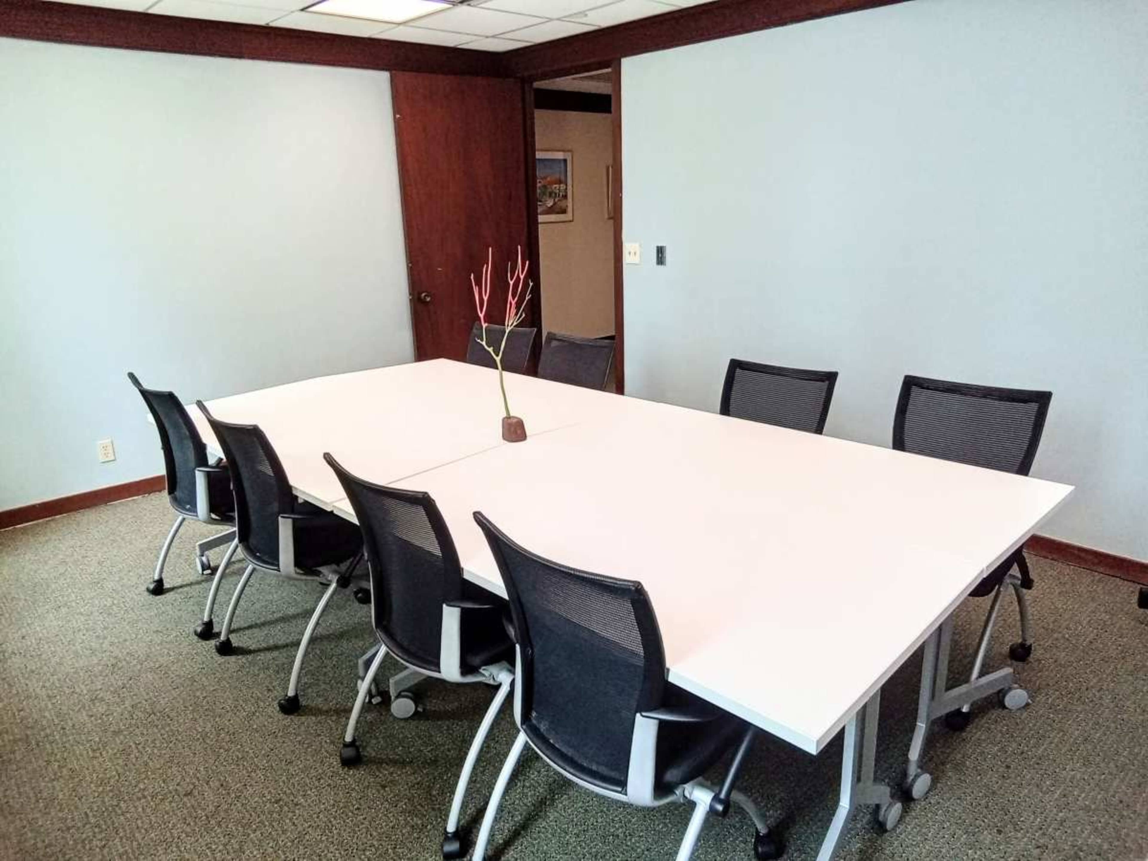 A long white table is surrounded by black mesh chairs in a conference room with a beige carpet and light blue walls.
