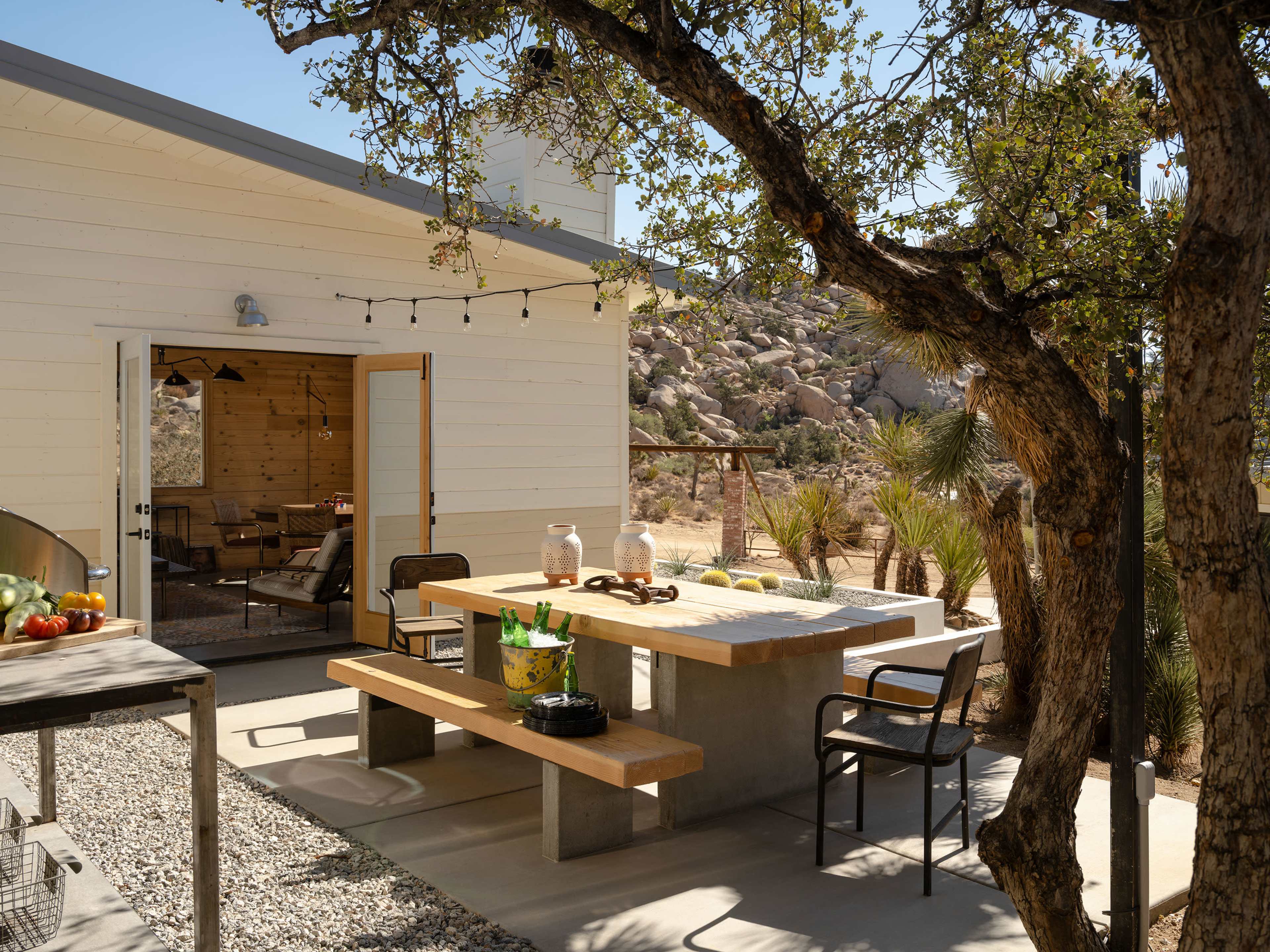 A modern outdoor dining area features a concrete table with wooden benches, surrounded by desert vegetation and a view of a rustic building.