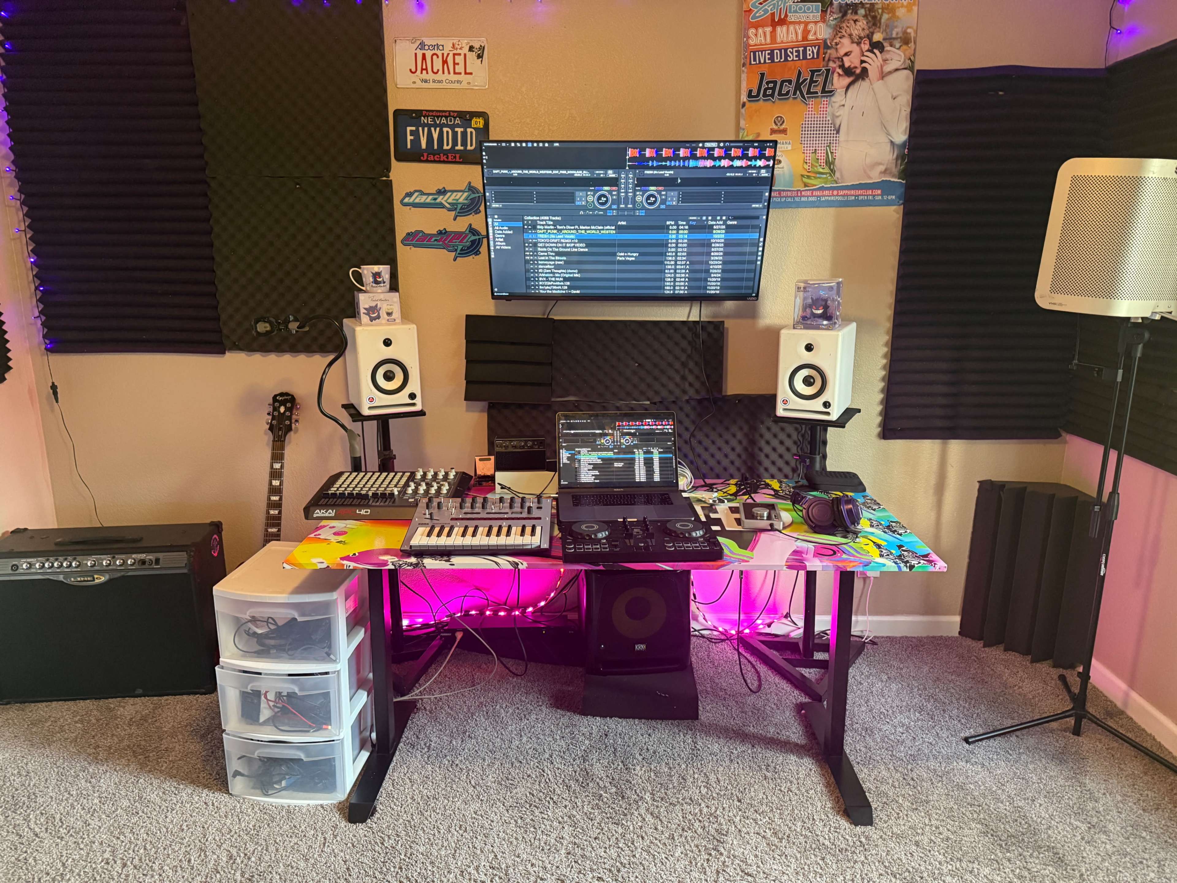 A home music production setup features a computer, audio speakers, a MIDI controller, and a DJ controller on a desk, with soundproof foam panels and colorful LED lighting in the background.