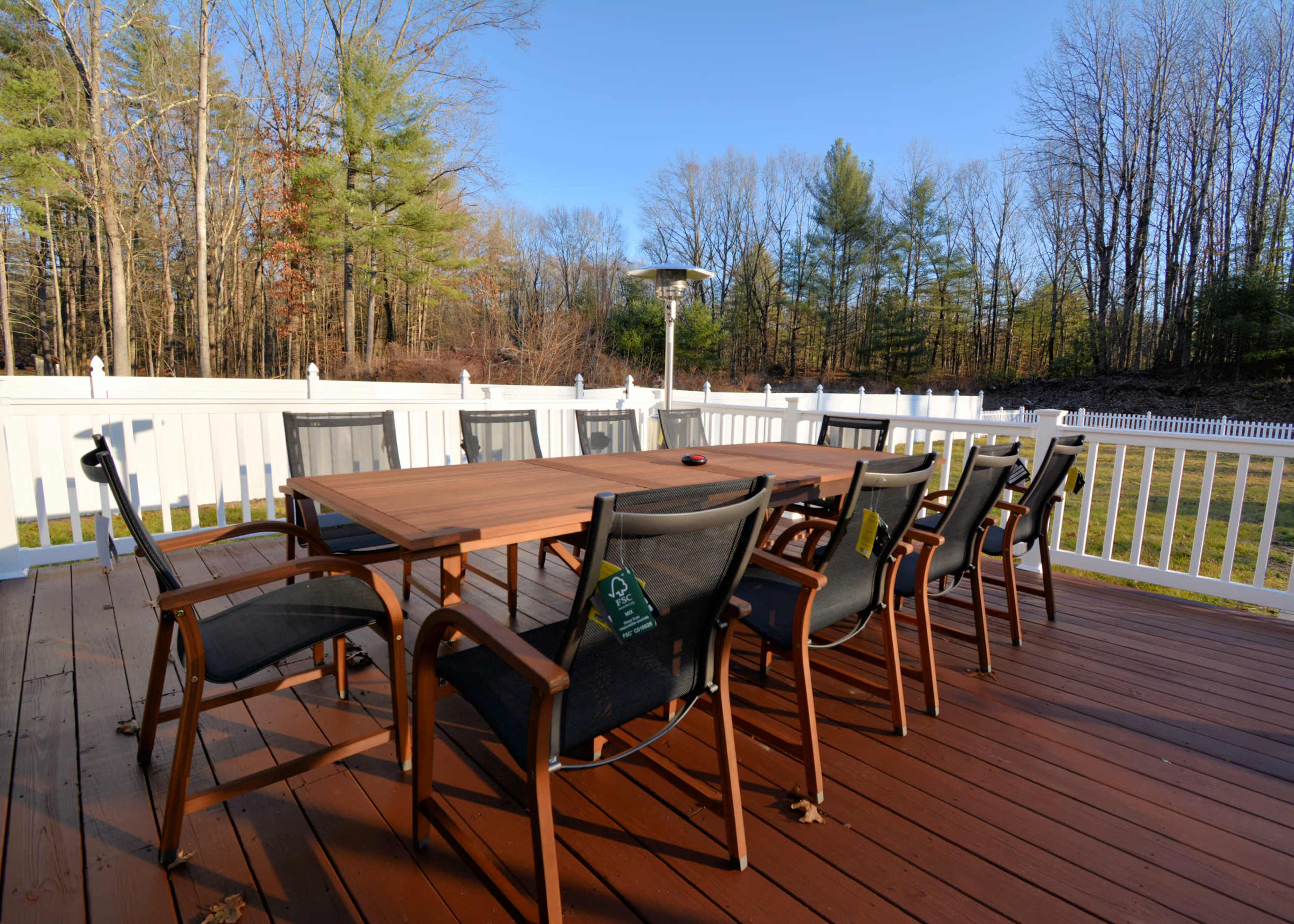 A spacious outdoor patio features a wooden dining table surrounded by several black chairs and a standing heater, set against a backdrop of trees and a white fence.