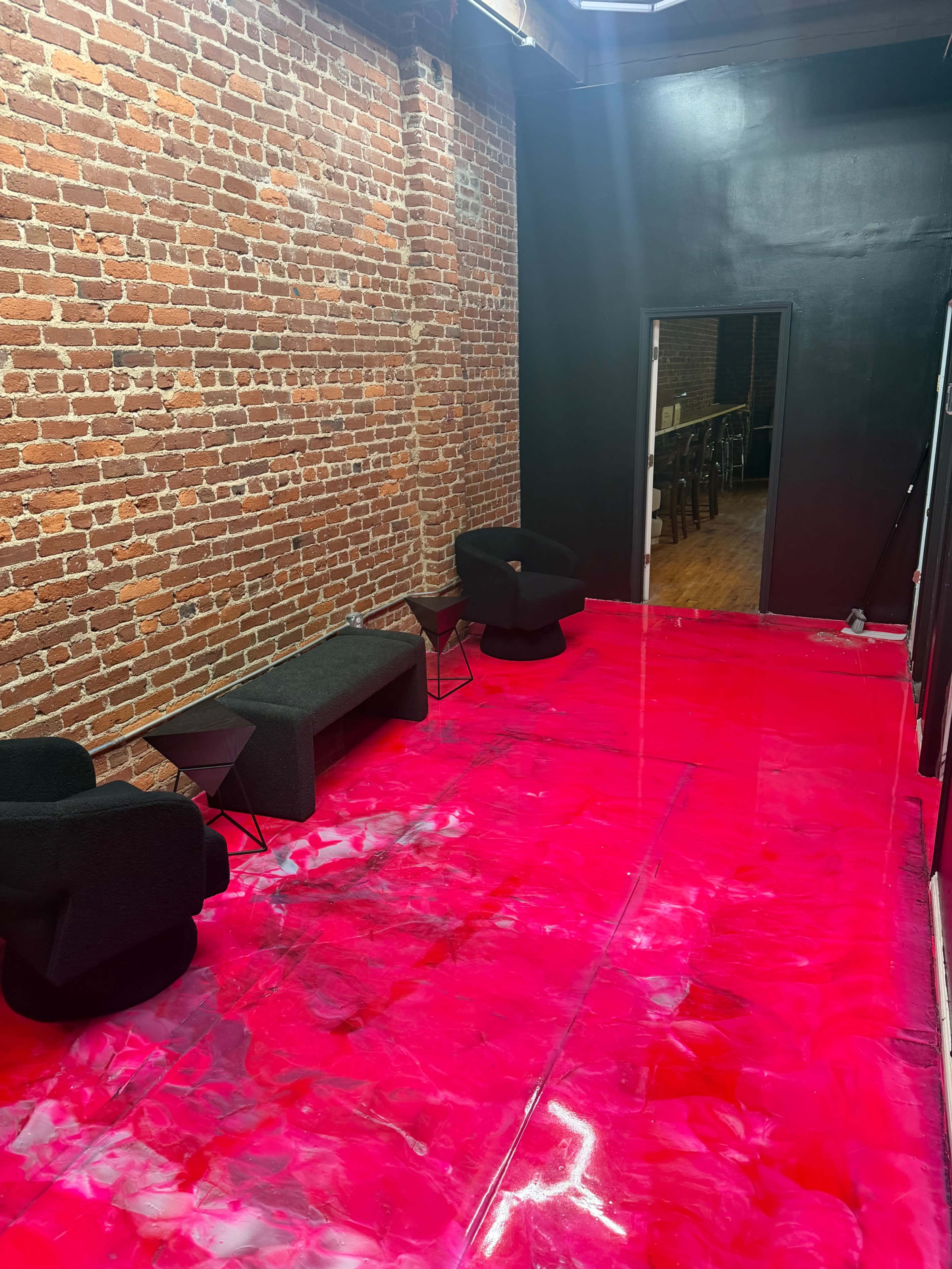 The image shows a room with exposed brick walls, featuring a bright pink floor and minimal furniture, including two black chairs and a long table.