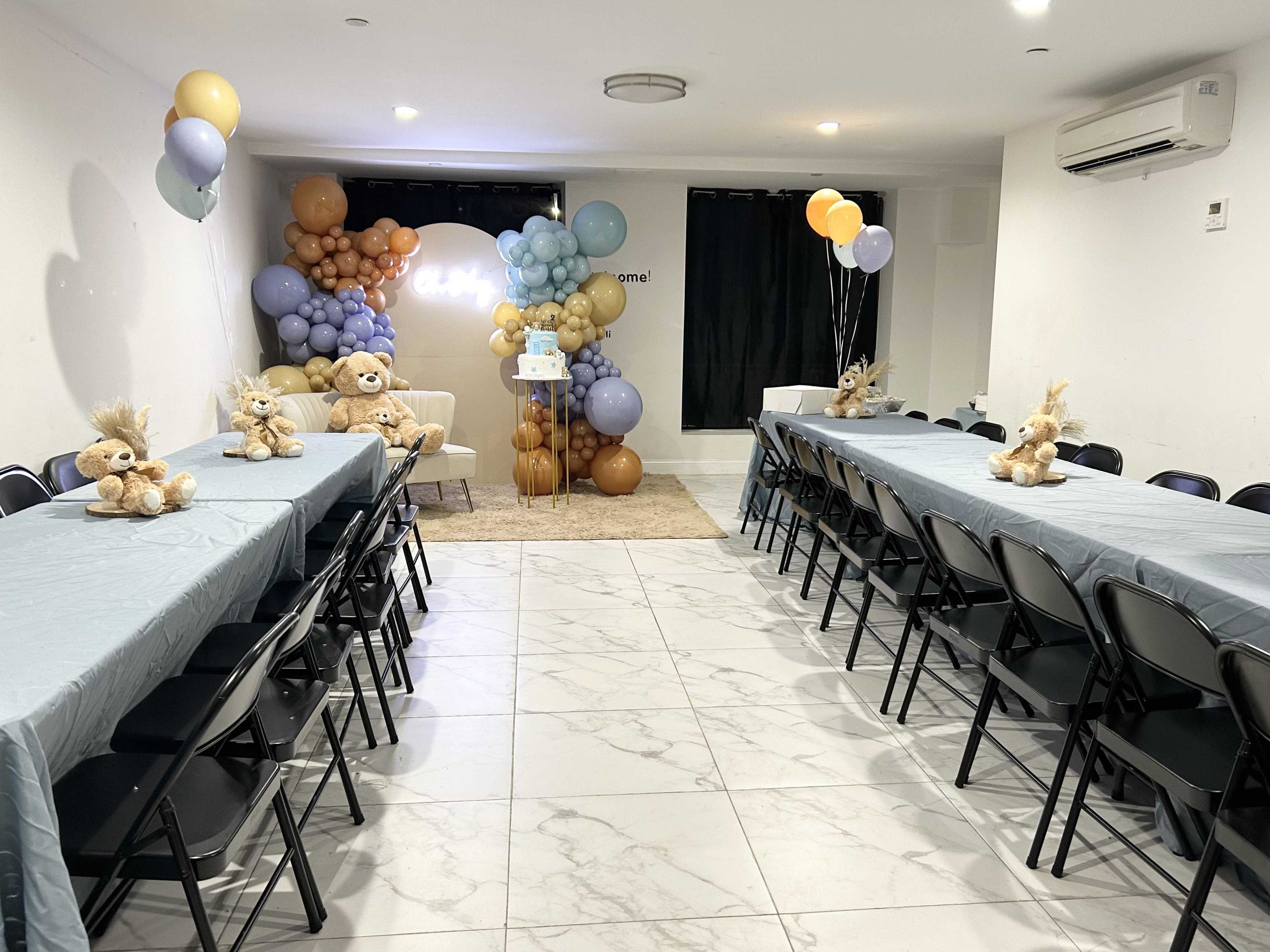 A spacious room is arranged for a party, featuring two long tables covered with light blue tablecloths, teddy bears on each table, and a backdrop of balloon decorations in various colors.