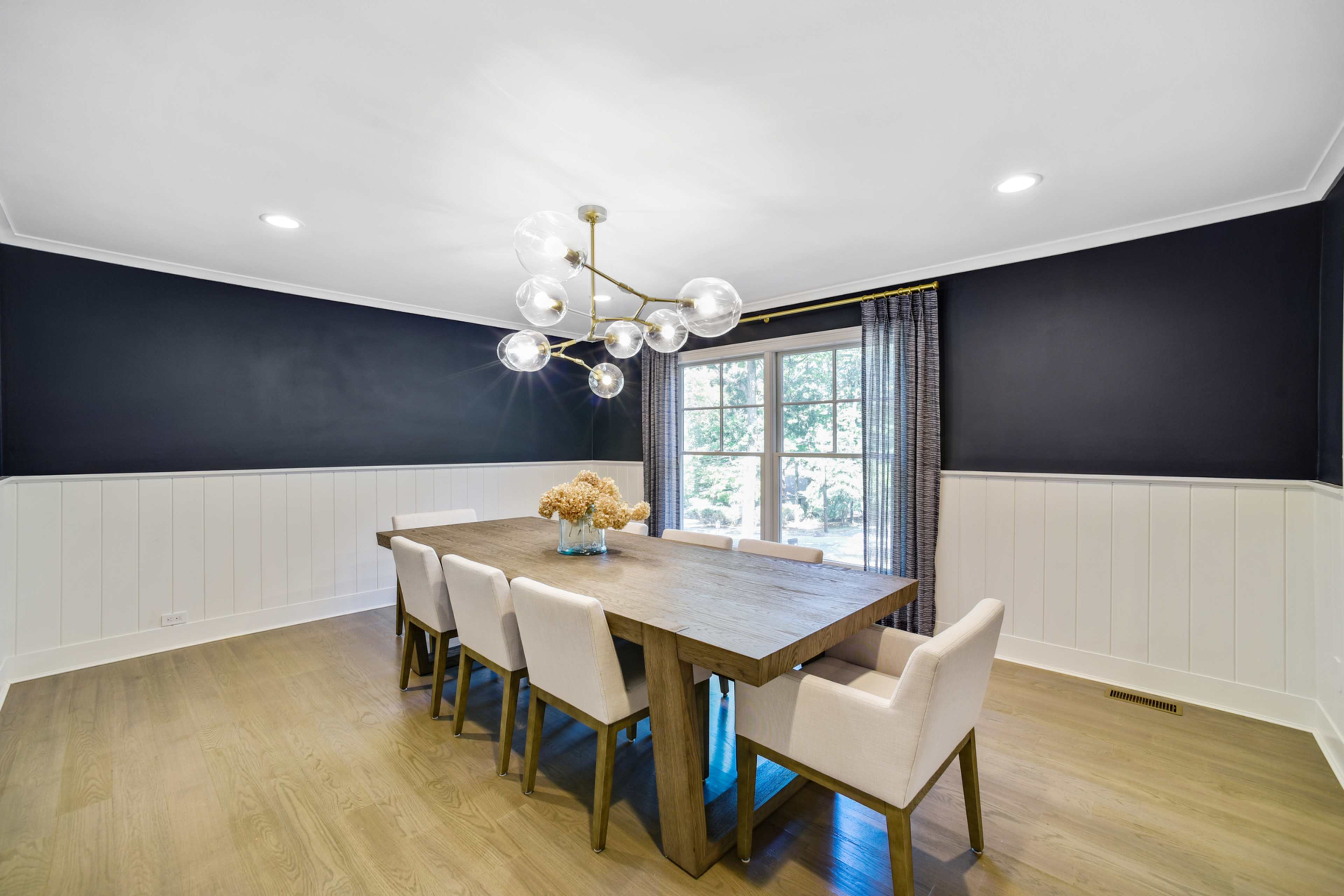 A wooden dining table with six chairs is positioned in a well-lit room featuring dark walls and a chandelier.