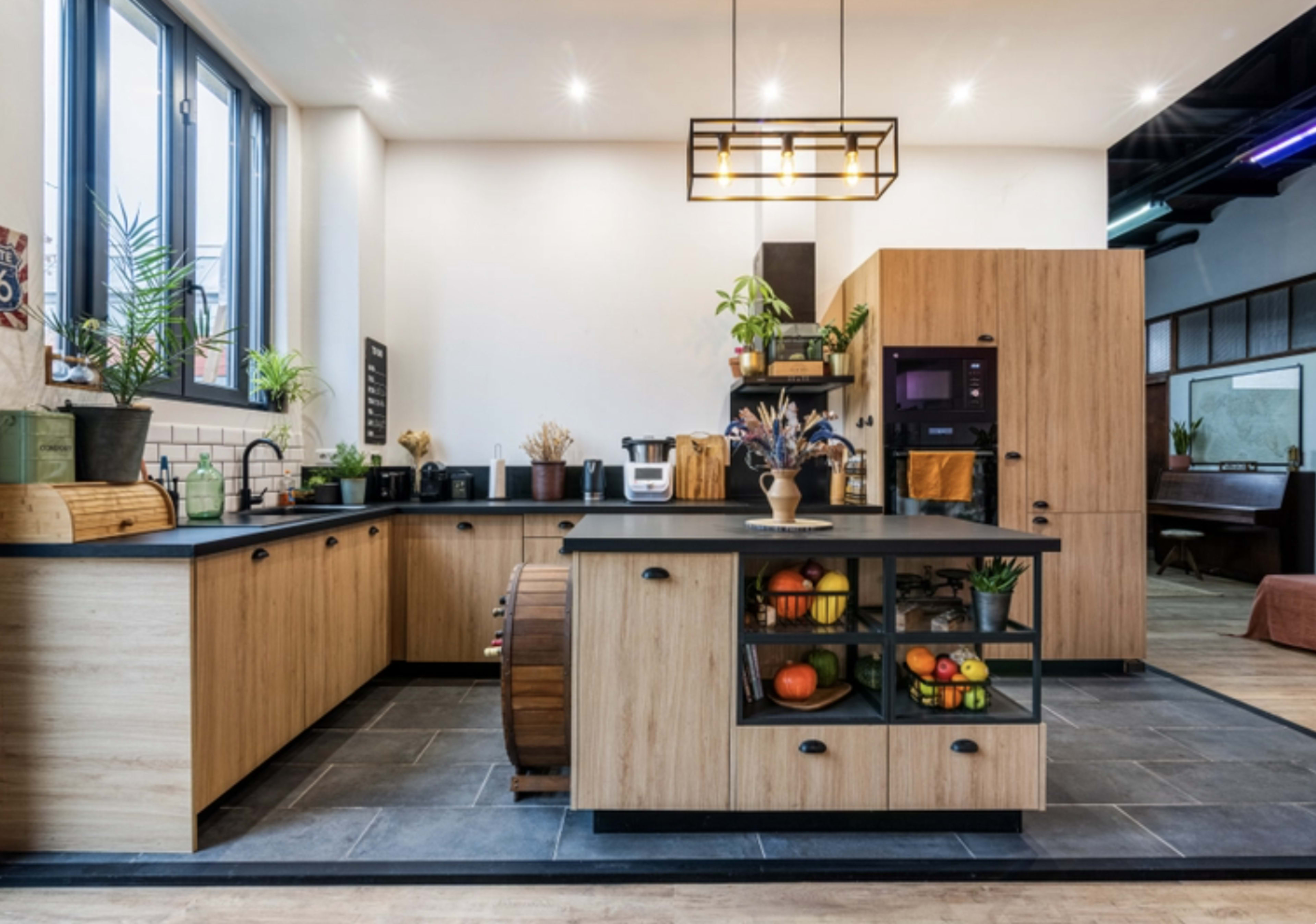 A modern kitchen featuring wooden cabinetry, a large island with black countertops, and various plants and fruits displayed throughout the space.