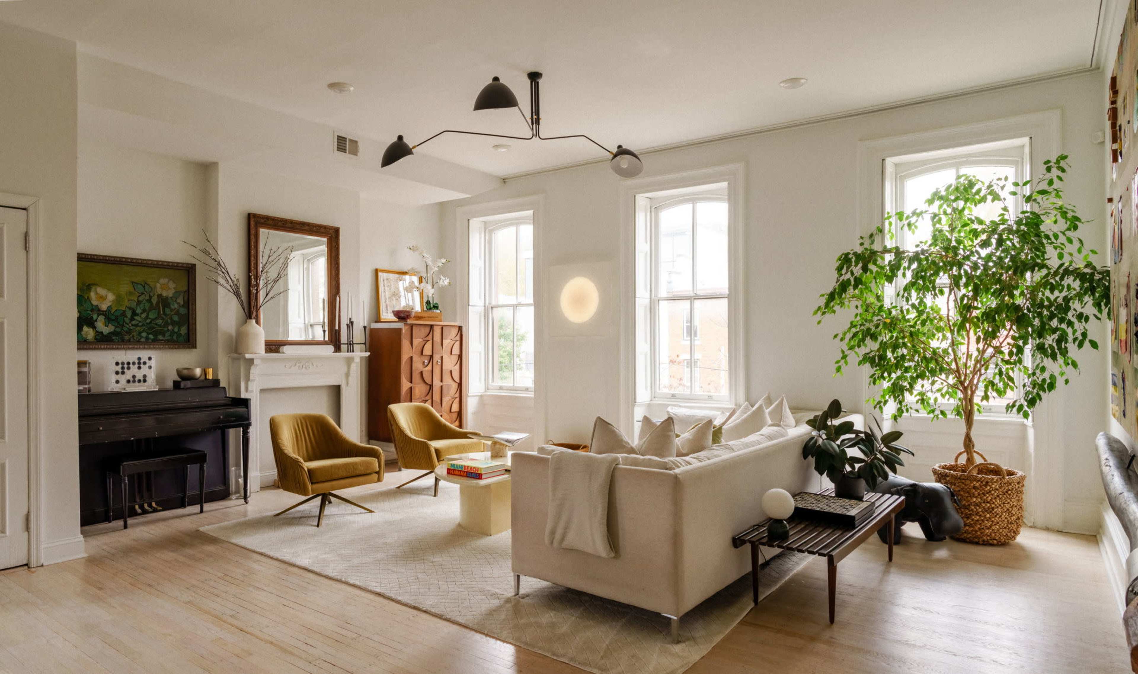 The image shows a bright living room featuring a sofa, two chairs, a coffee table, and a piano, with large windows allowing natural light to enter and a decorative plant in the corner.