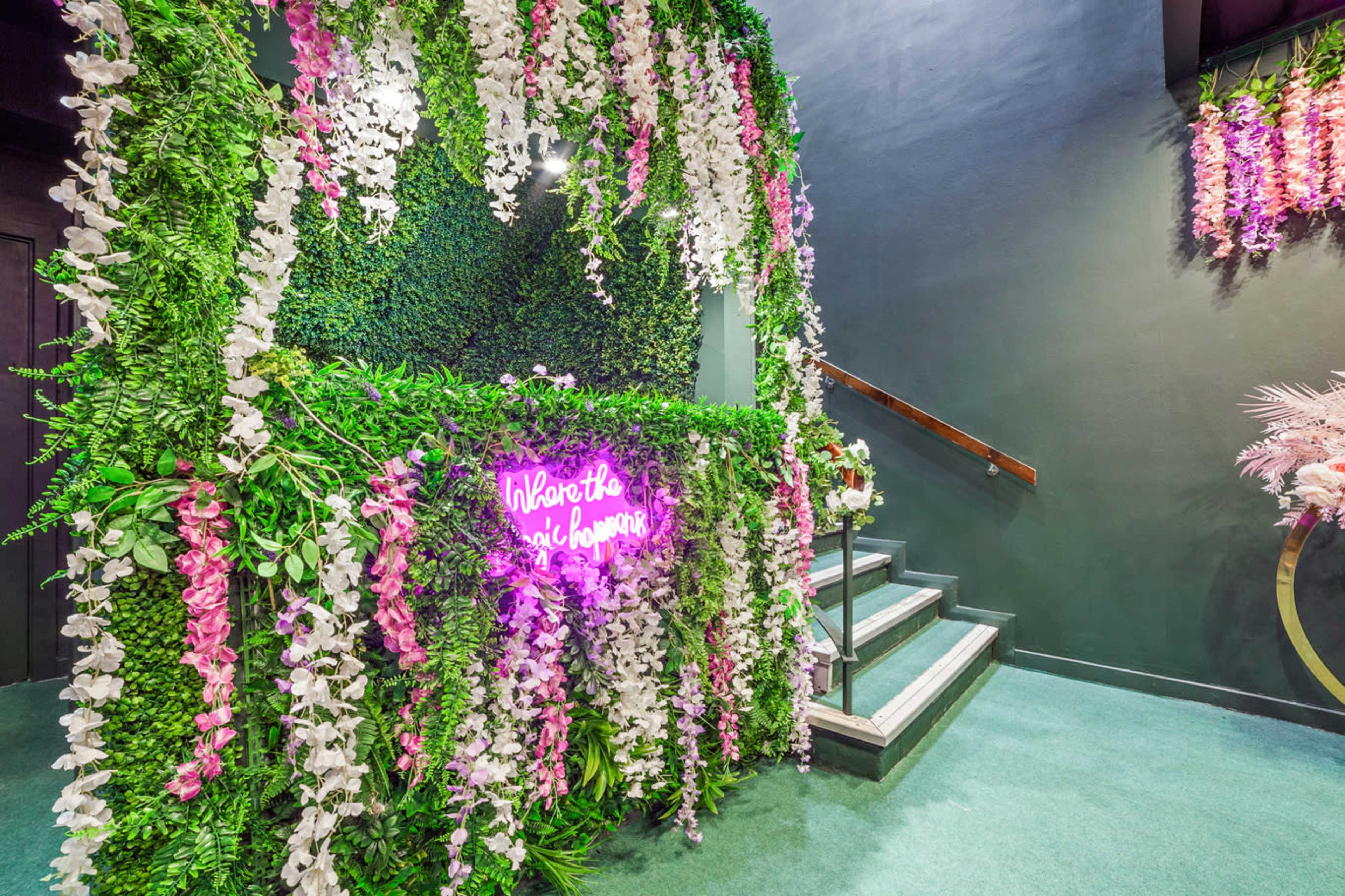 A floral-themed entrance area with decorative greenery and bright pink neon signage, leading up a set of stairs.