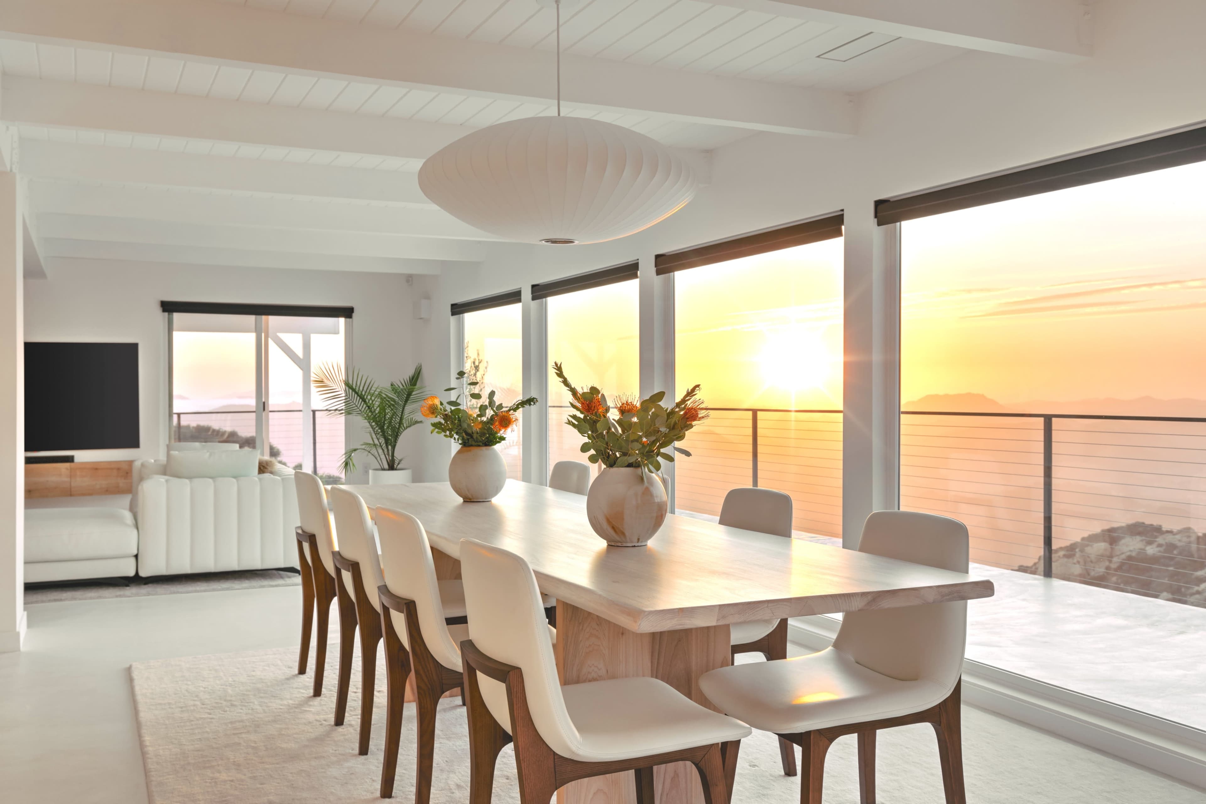 A modern dining room features a long wooden table set for eight, with vases of flowers, large windows framing a sunset view, and minimalist decor.