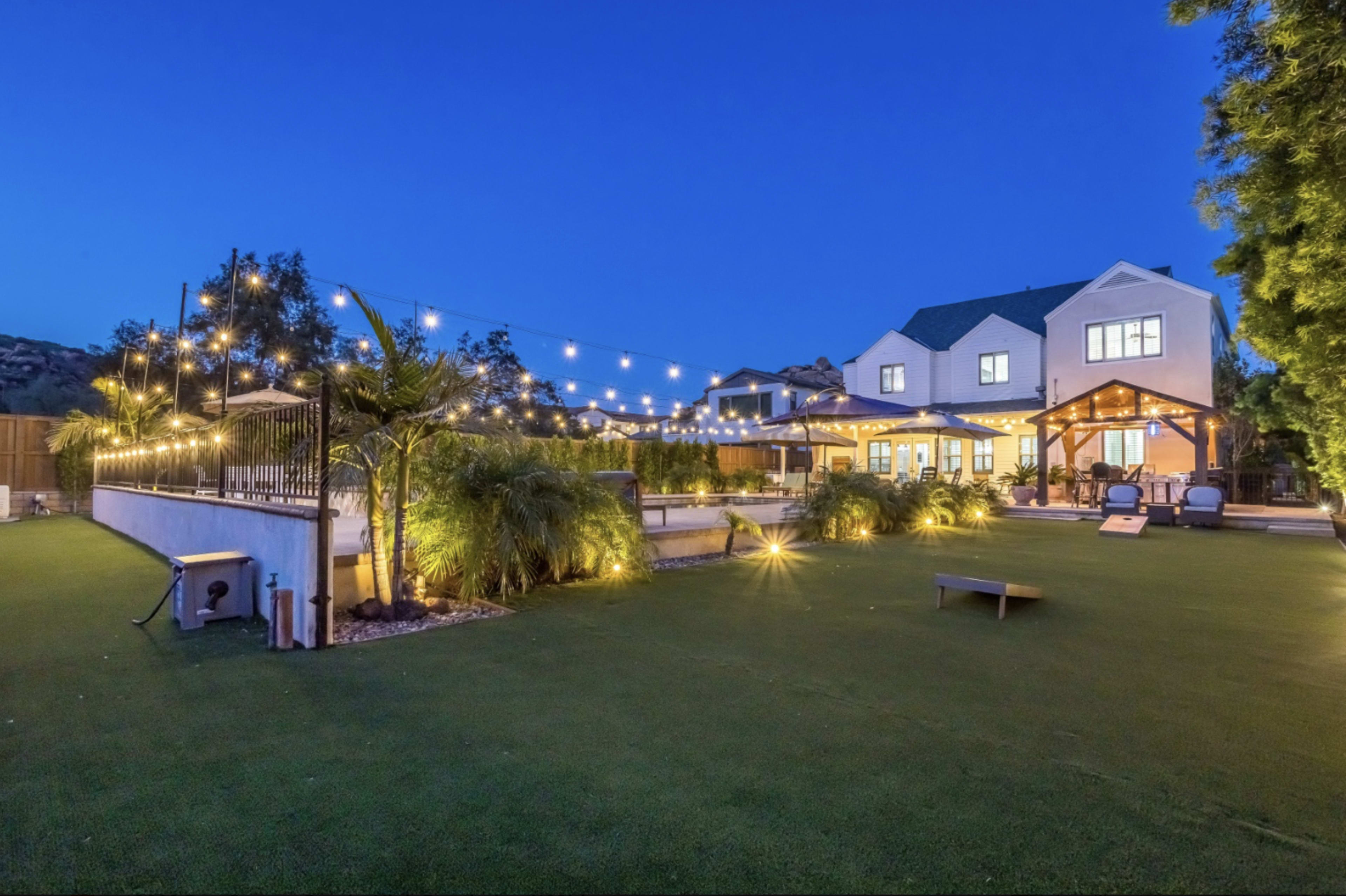 A backyard is illuminated by string lights and features a patio, seating area, and well-maintained landscaping, alongside a large house.