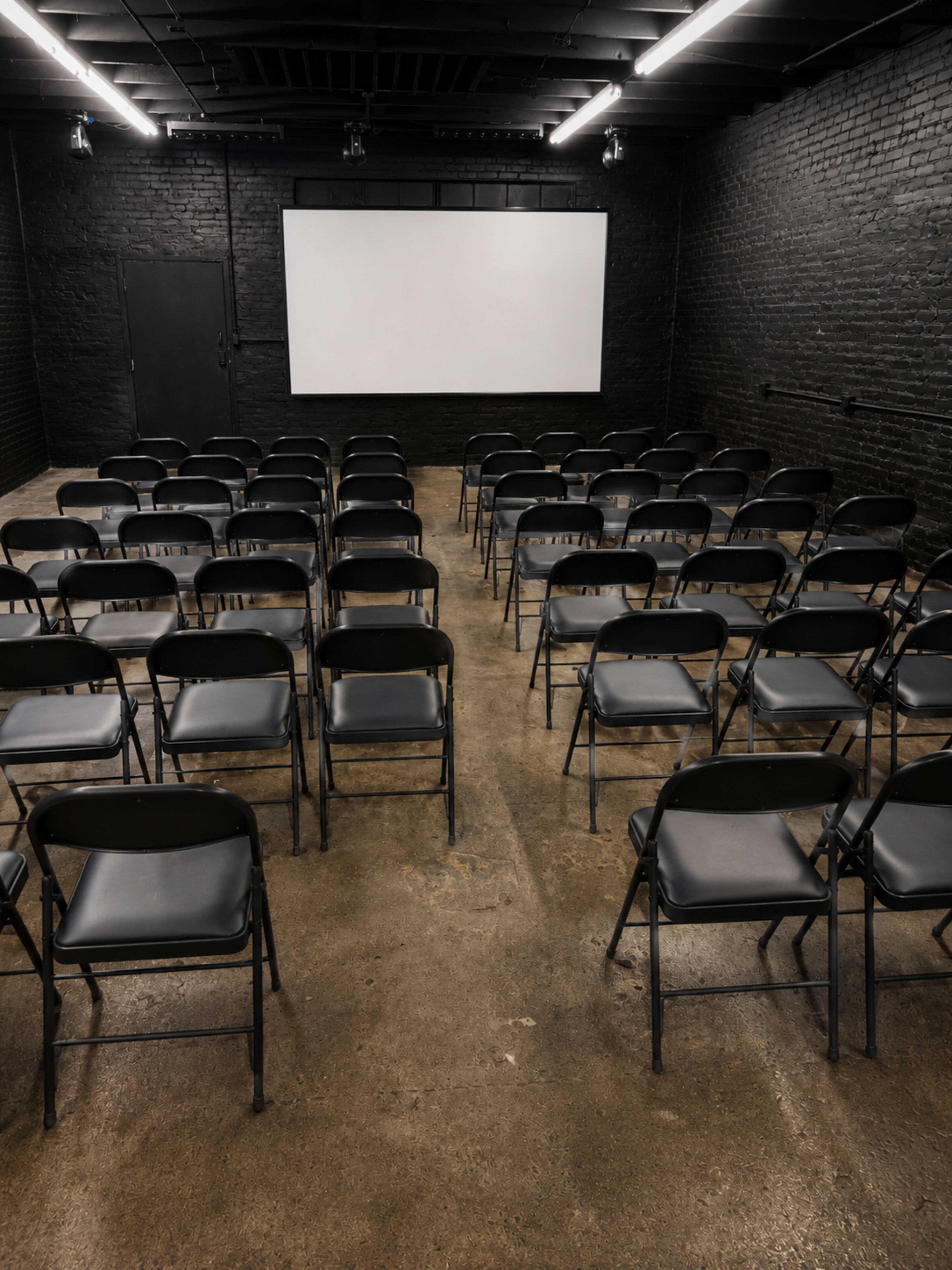 The image shows a room with rows of black folding chairs facing a blank white screen on a brick wall.