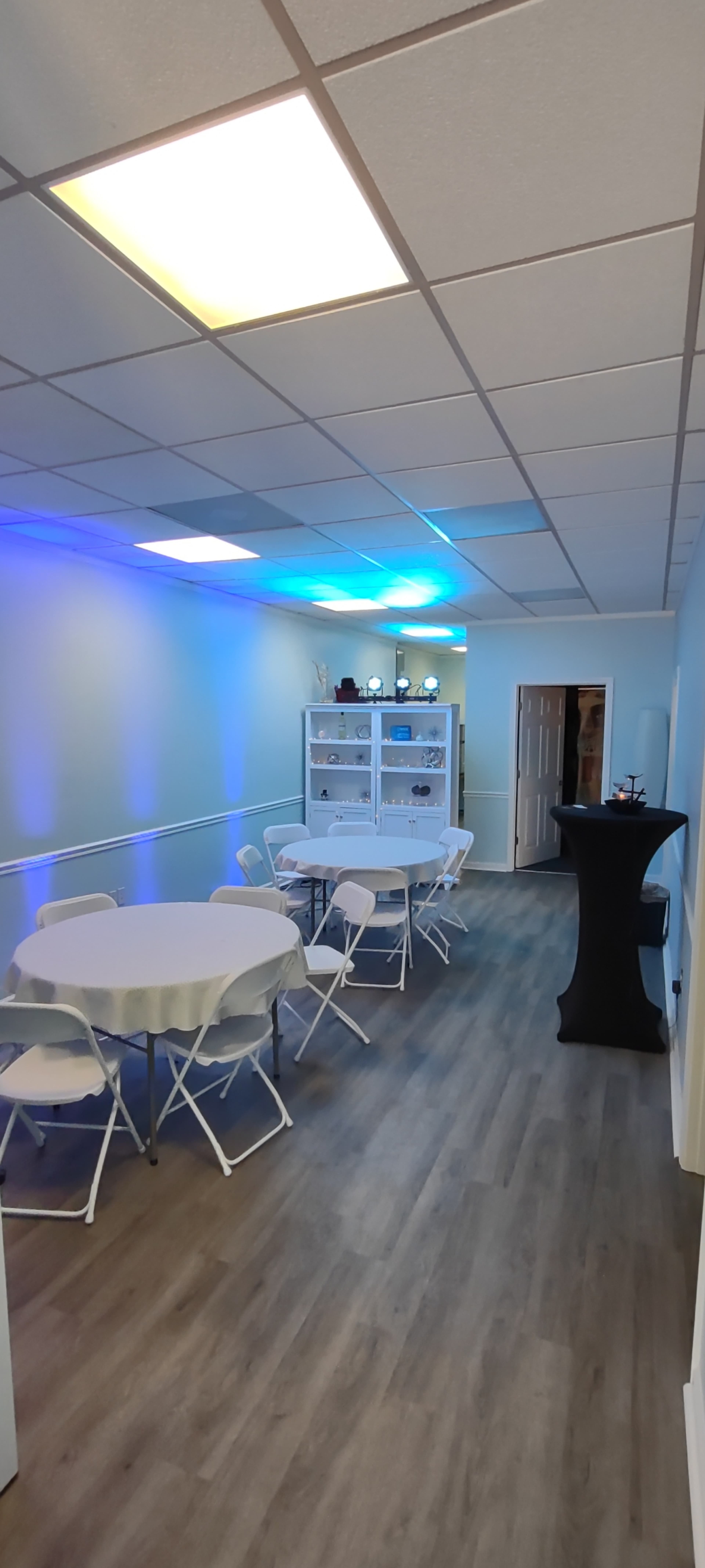 The image shows a narrow room with white walls, featuring a round table with chairs, a black high table, and shelves displaying various items, all illuminated by blue lighting.