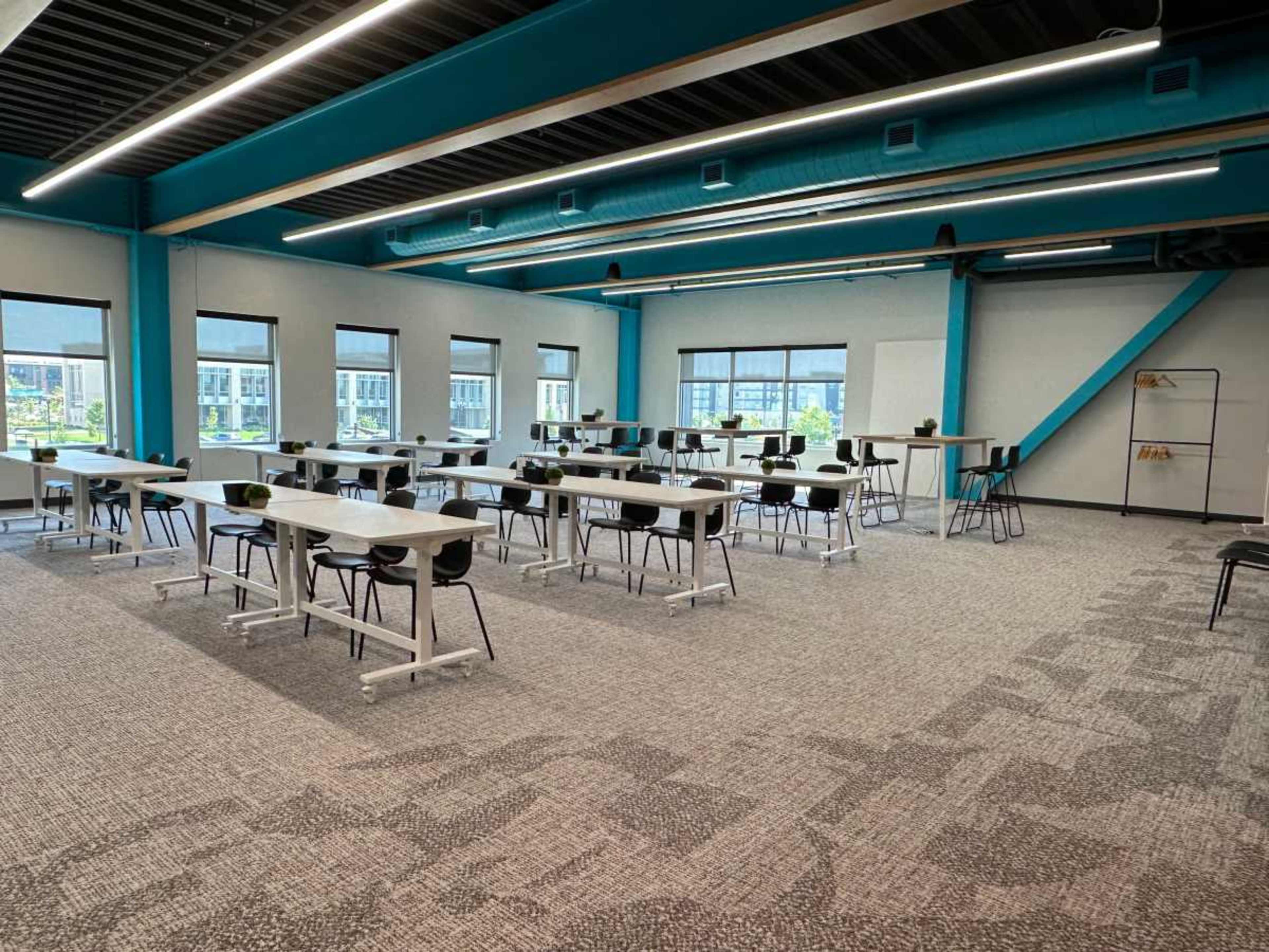 The image shows a spacious, modern room arranged with multiple tables and chairs, featuring large windows that allow natural light to illuminate the area.