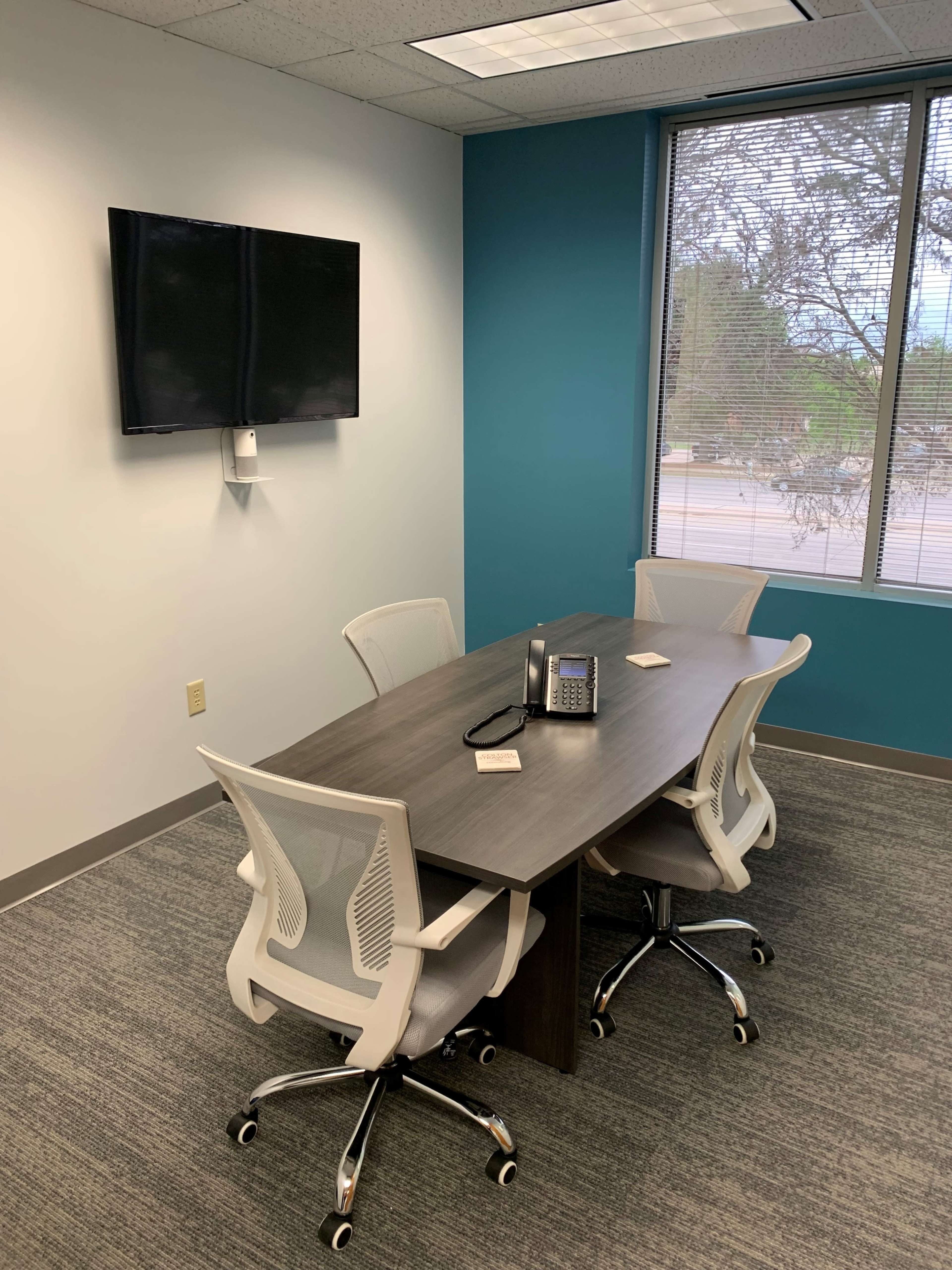 A small conference room with a rectangular table, four rolling chairs, a television mounted on the wall, and a window allowing natural light.