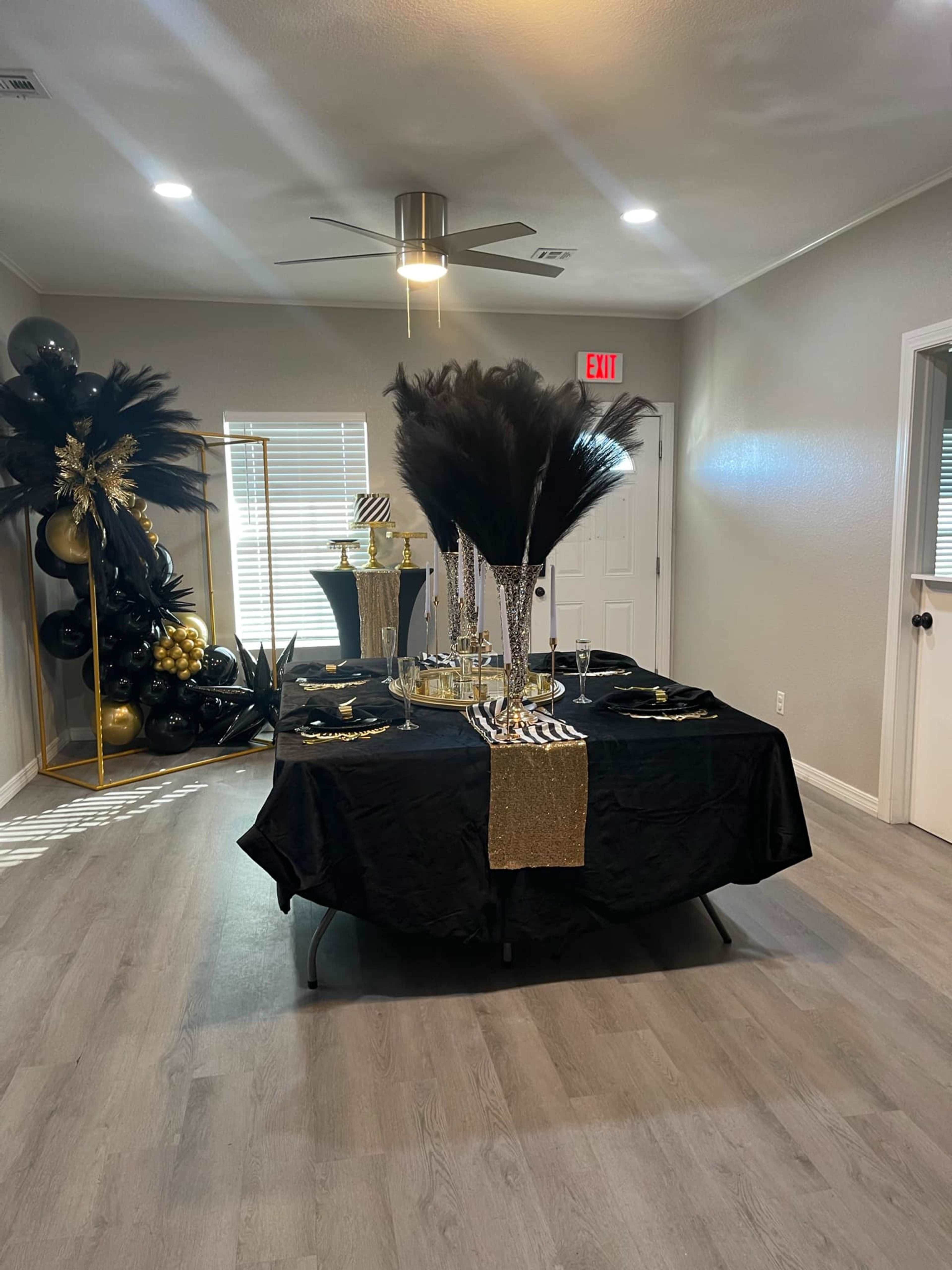 The image shows a decorated event space with a black table adorned with a gold and black centerpiece, surrounded by balloons and elegant table settings.
