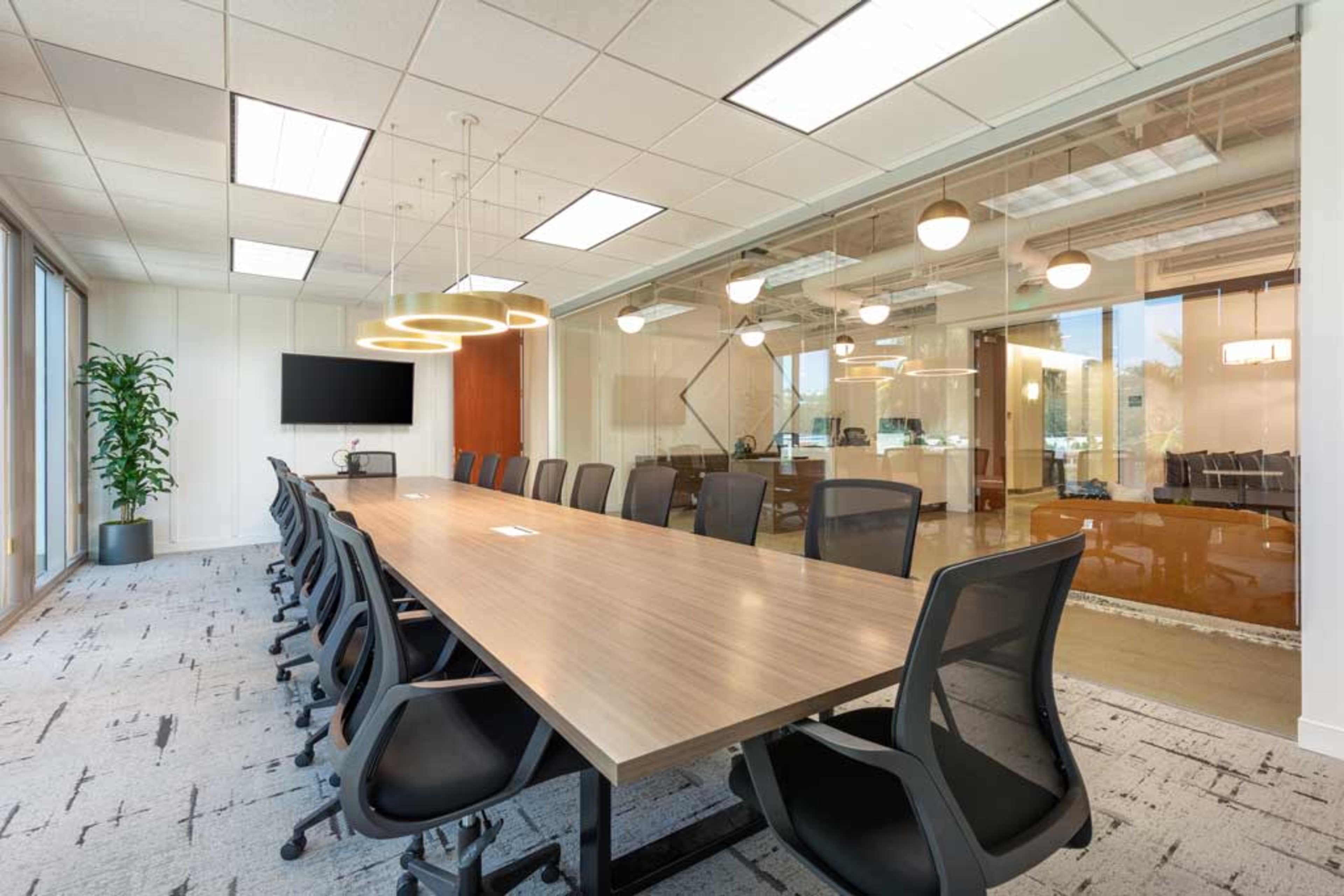 A large conference room with a long rectangular table, multiple black office chairs, and glass walls separating it from another office space.