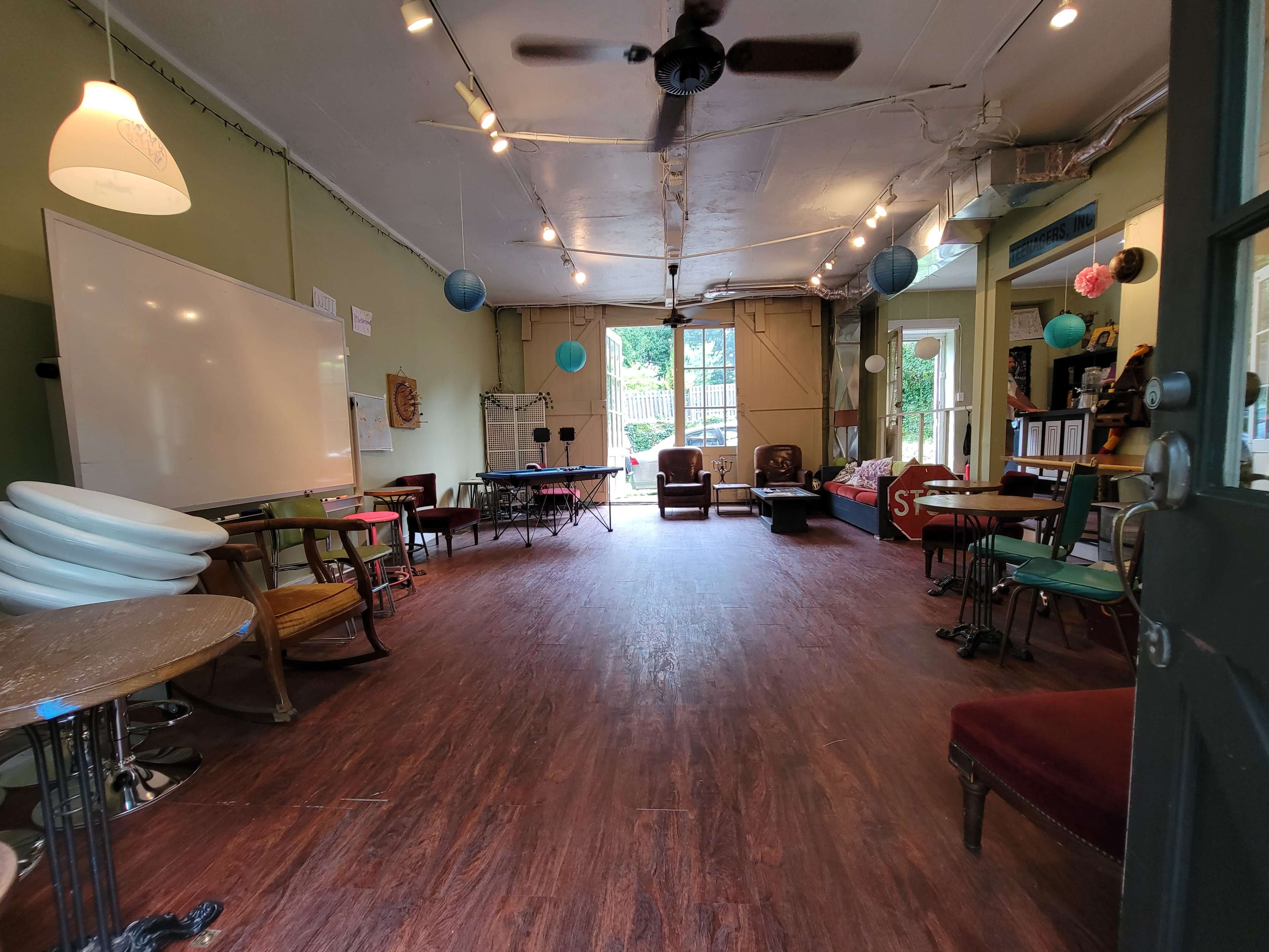 The image shows a spacious, empty room with wooden floors and various seating arrangements, including chairs and tables, leading to a large doorway that opens to an outdoor area.