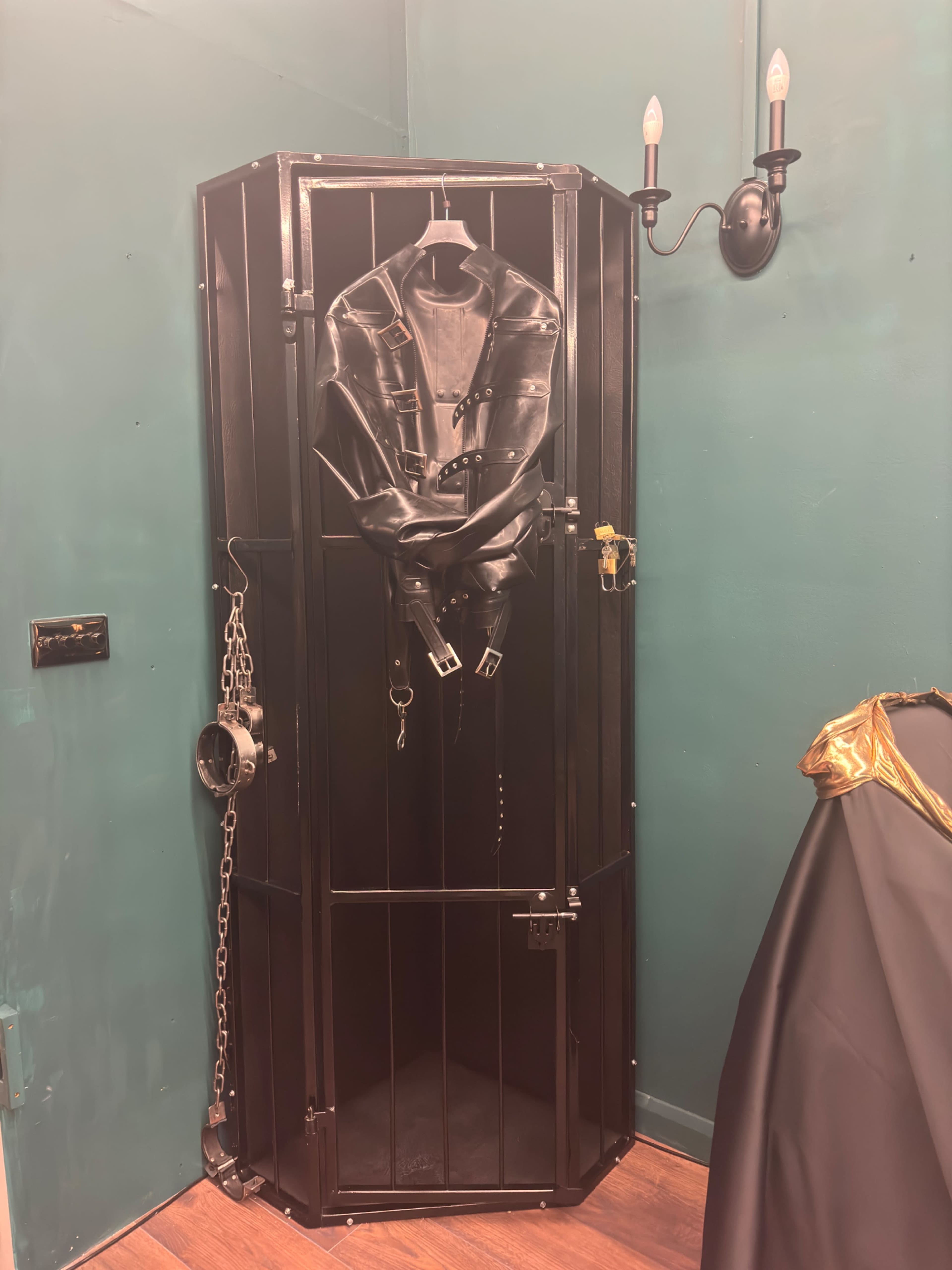 The image shows a black metal cage in the corner of a room, containing a leather outfit hanging from a hook and accompanied by a pair of handcuffs and a chain.
