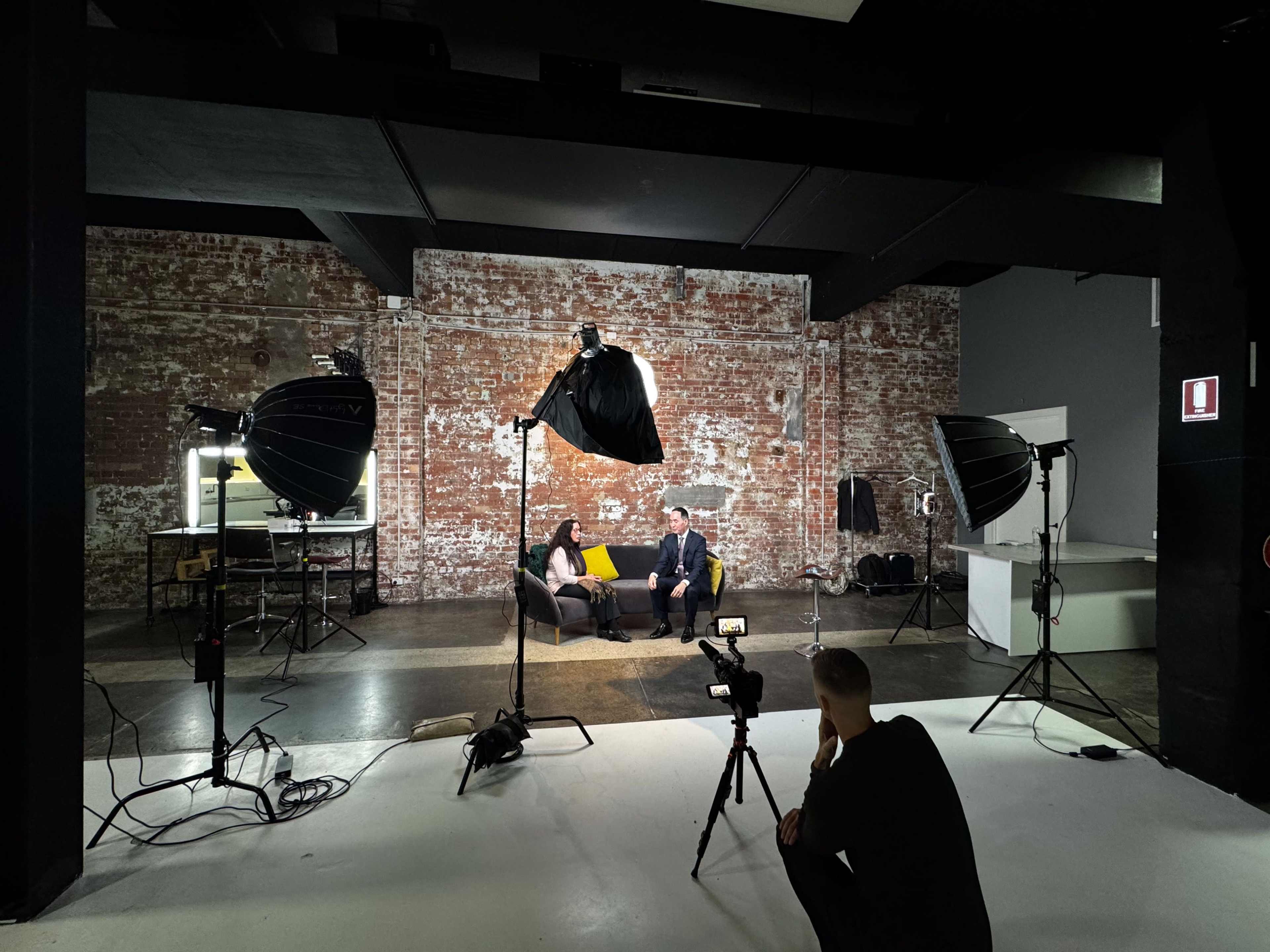 A man and a woman are seated on a couch in a dimly lit studio, surrounded by photography lighting equipment and a camera operator in the foreground.