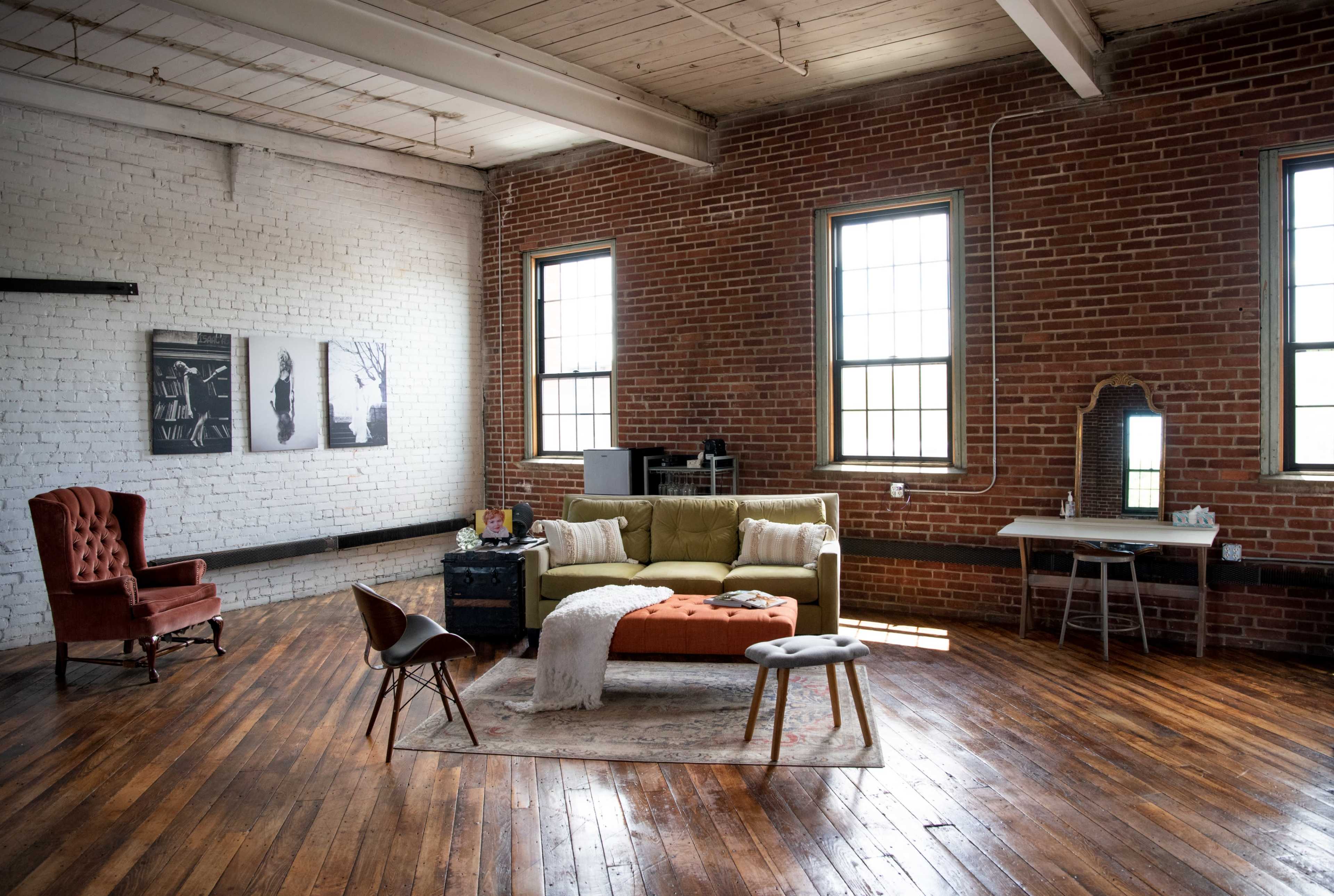 A spacious room with exposed brick walls, wooden floors, a green sofa, a rust-colored ottoman, and a small desk near two large windows.