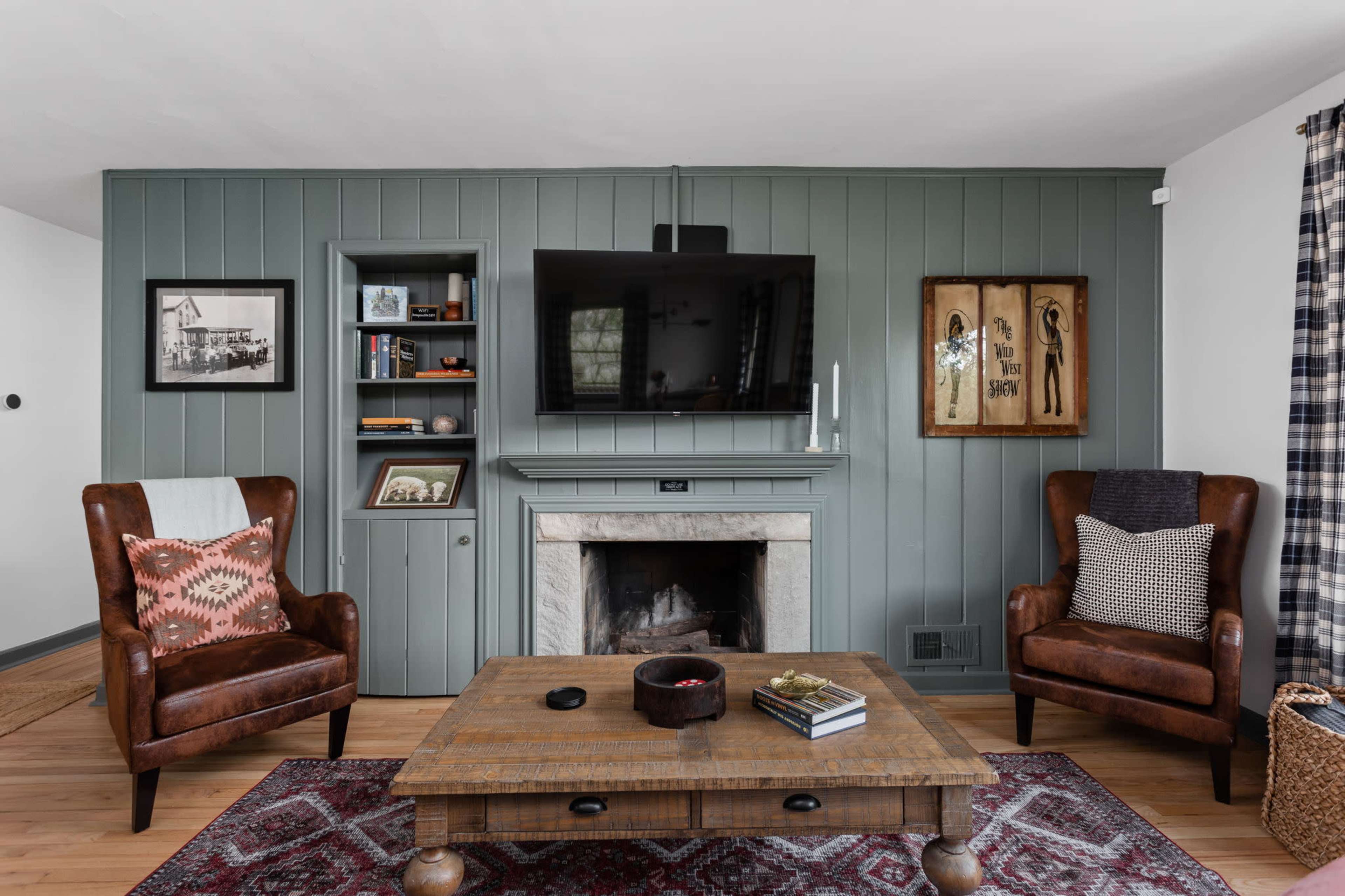 The living room features a wood-paneled wall, a mounted television above a fireplace, and two leather armchairs flanking a wooden coffee table.