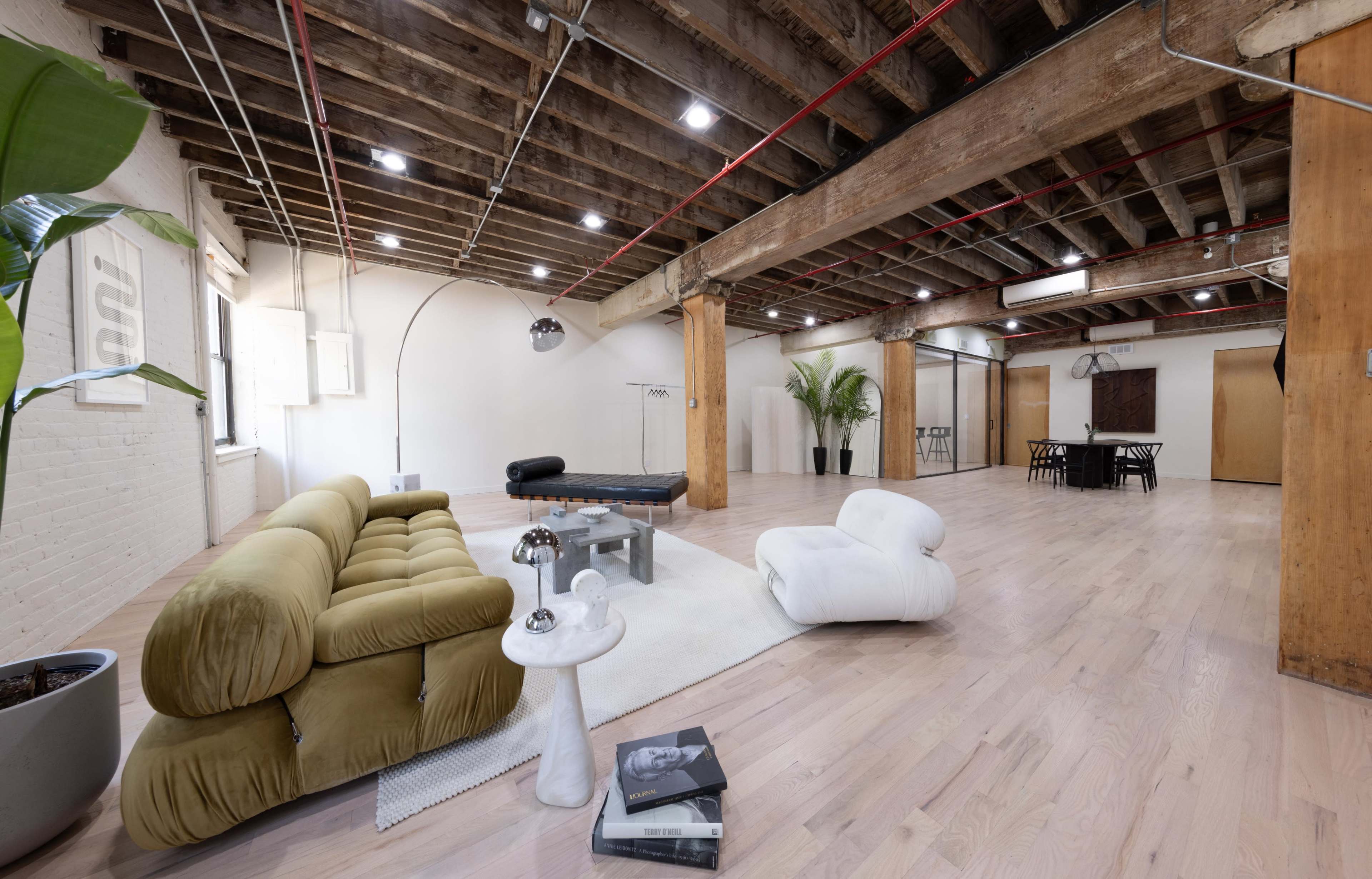 A spacious, industrial-style interior with wood beams, a light wooden floor, and minimalist furnishings including a green sofa, a coffee table, and a dining area.