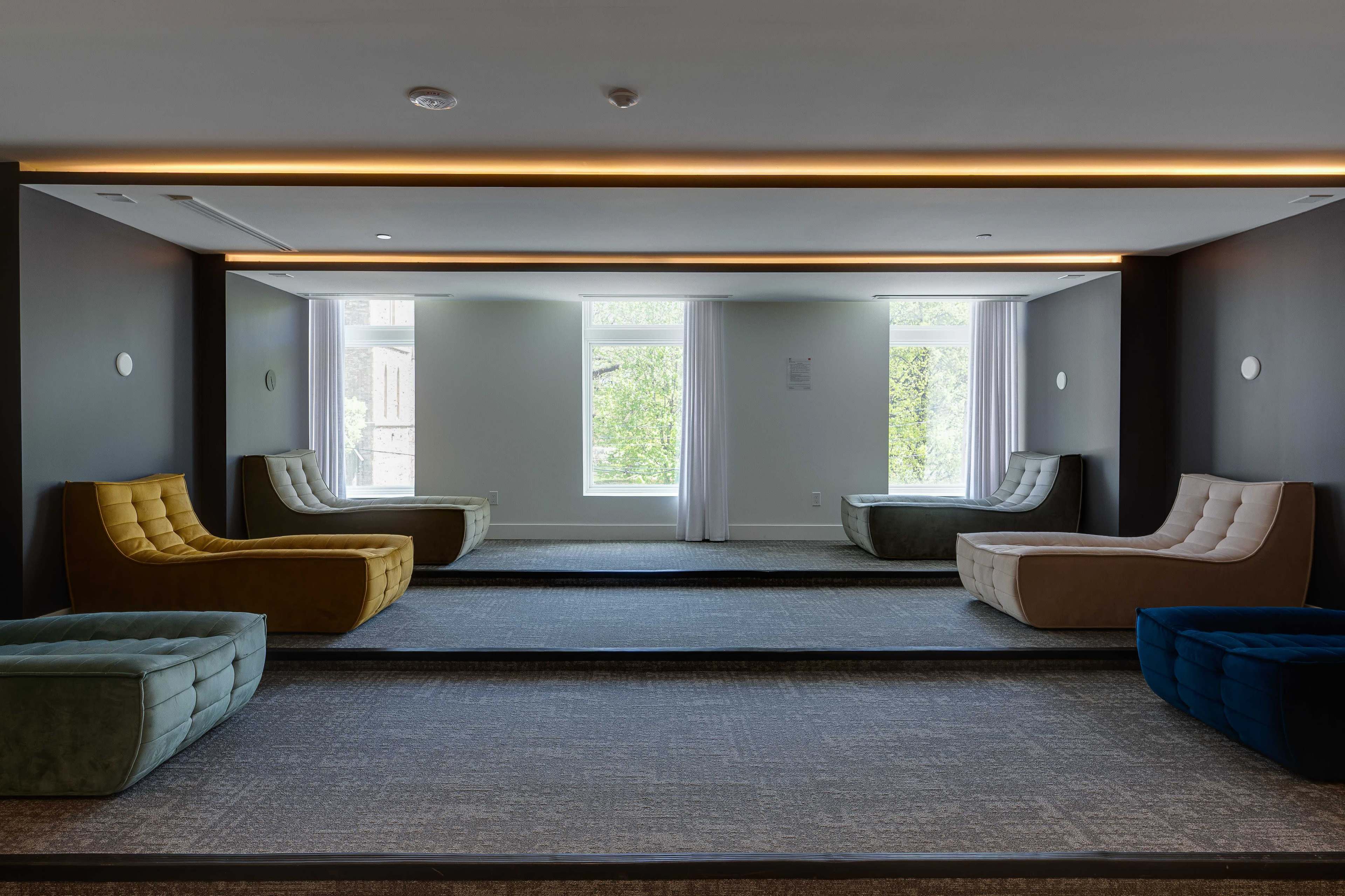 The image shows a modern lounge area featuring several upholstered curved benches in various colors, a light gray carpet, and large windows allowing natural light.
