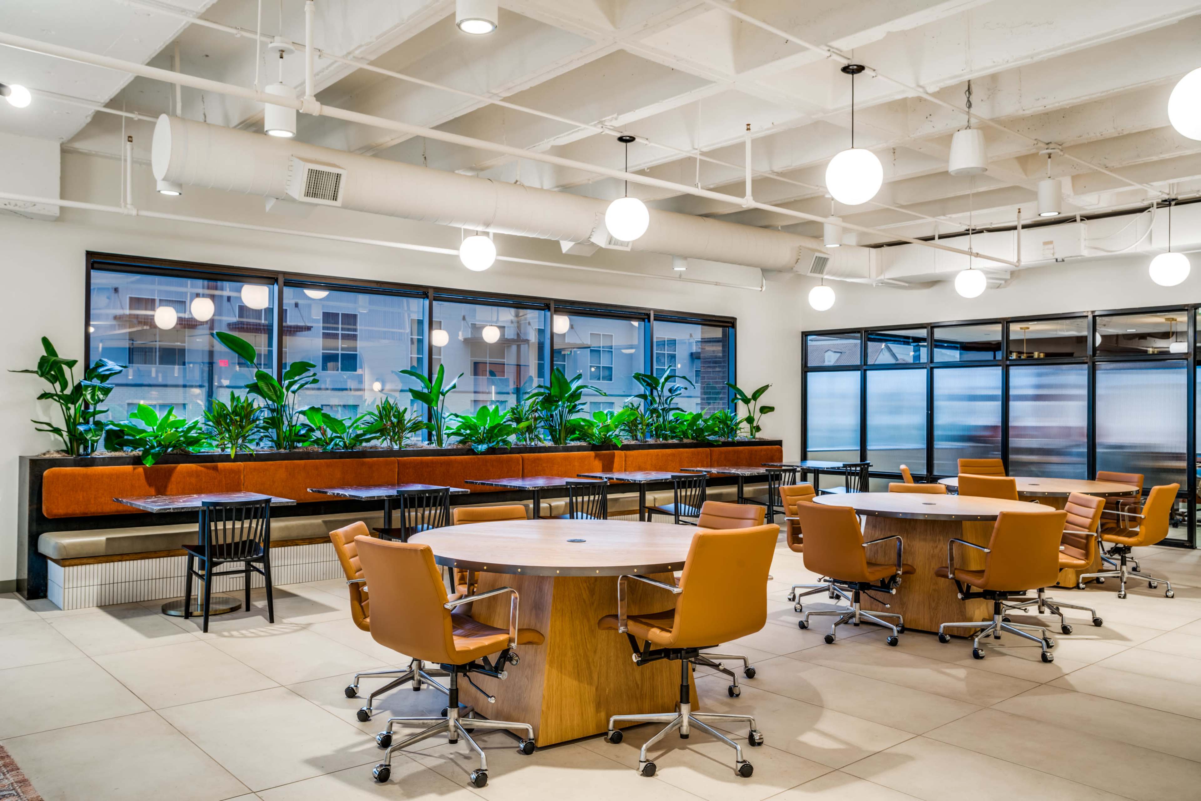 The image shows a modern office space featuring round tables surrounded by orange chairs and large windows with potted plants along the walls.