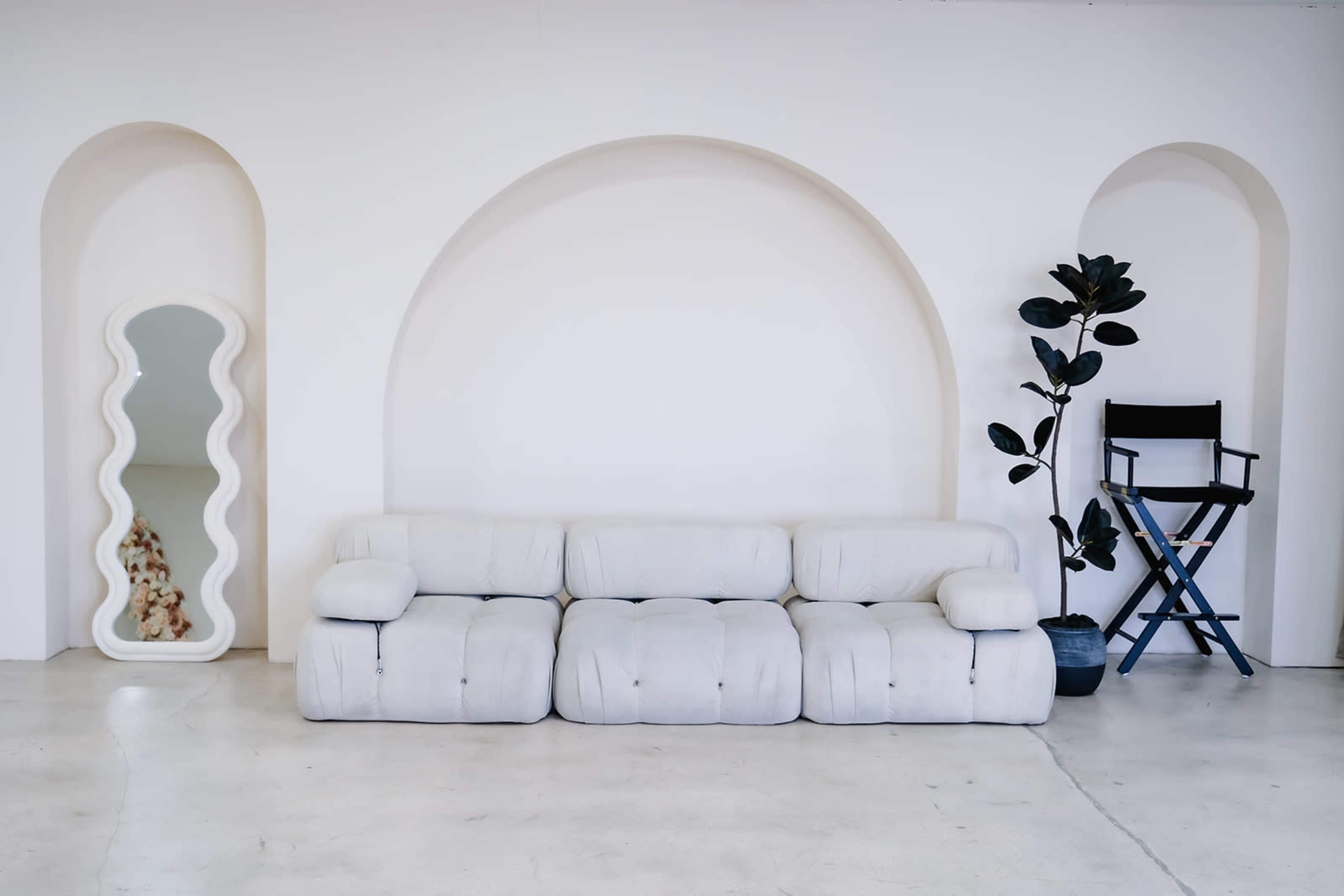 The image features a minimalist interior with a white, cushioned sectional sofa, a wavy mirror, a leafy plant in a pot, and a director's chair against a softly curved wall.
