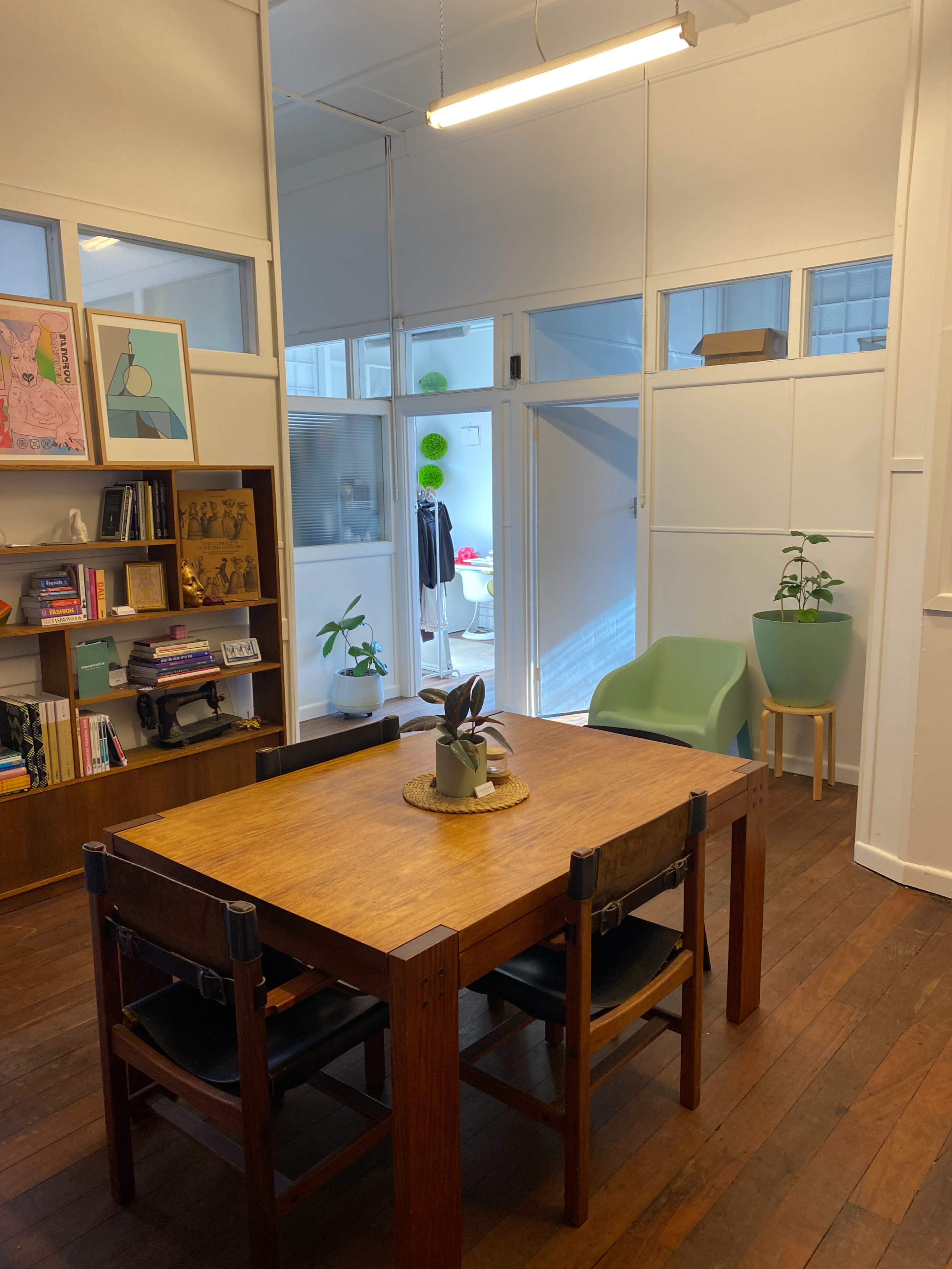 A wooden table with black chairs is positioned in a room featuring bookshelves, plants, and a green chair, illuminated by a hanging light.