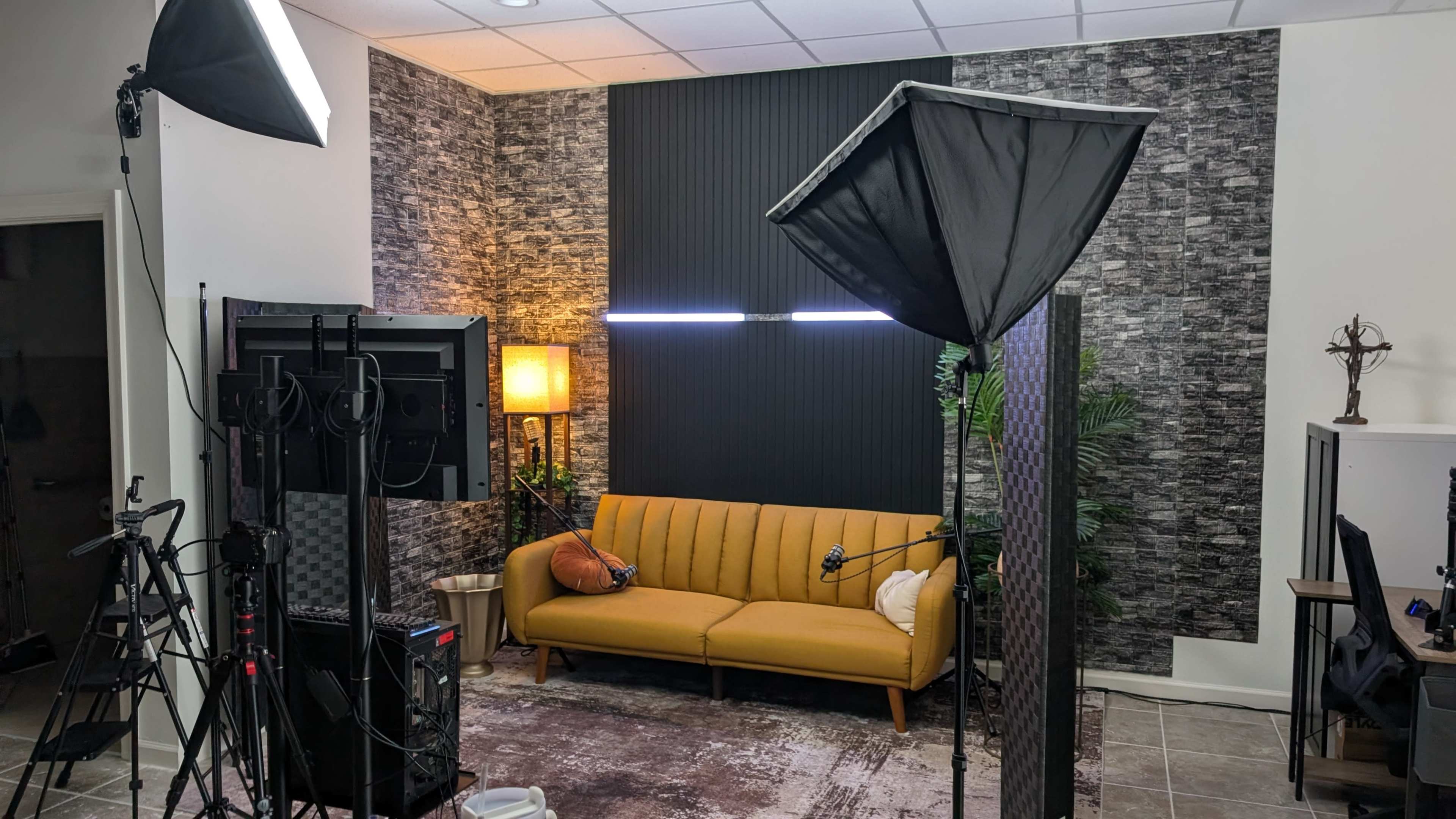 The image depicts a well-lit studio setup featuring a yellow sofa, lighting equipment, cameras, and a decorative backdrop with plants.