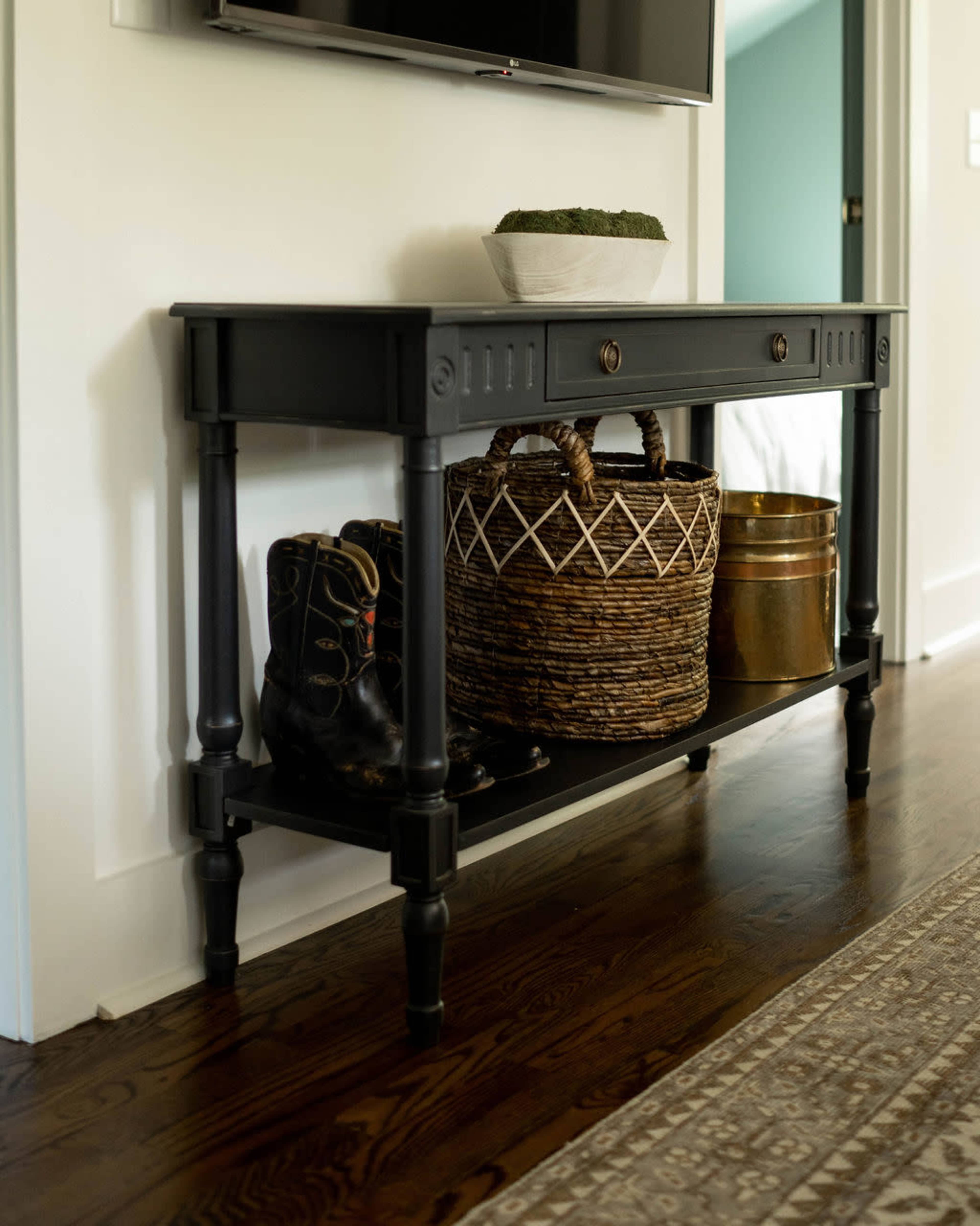 A dark wooden console table with a drawer sits against a wall, displaying a decorative basket and a metal container, along with a pair of black boots.