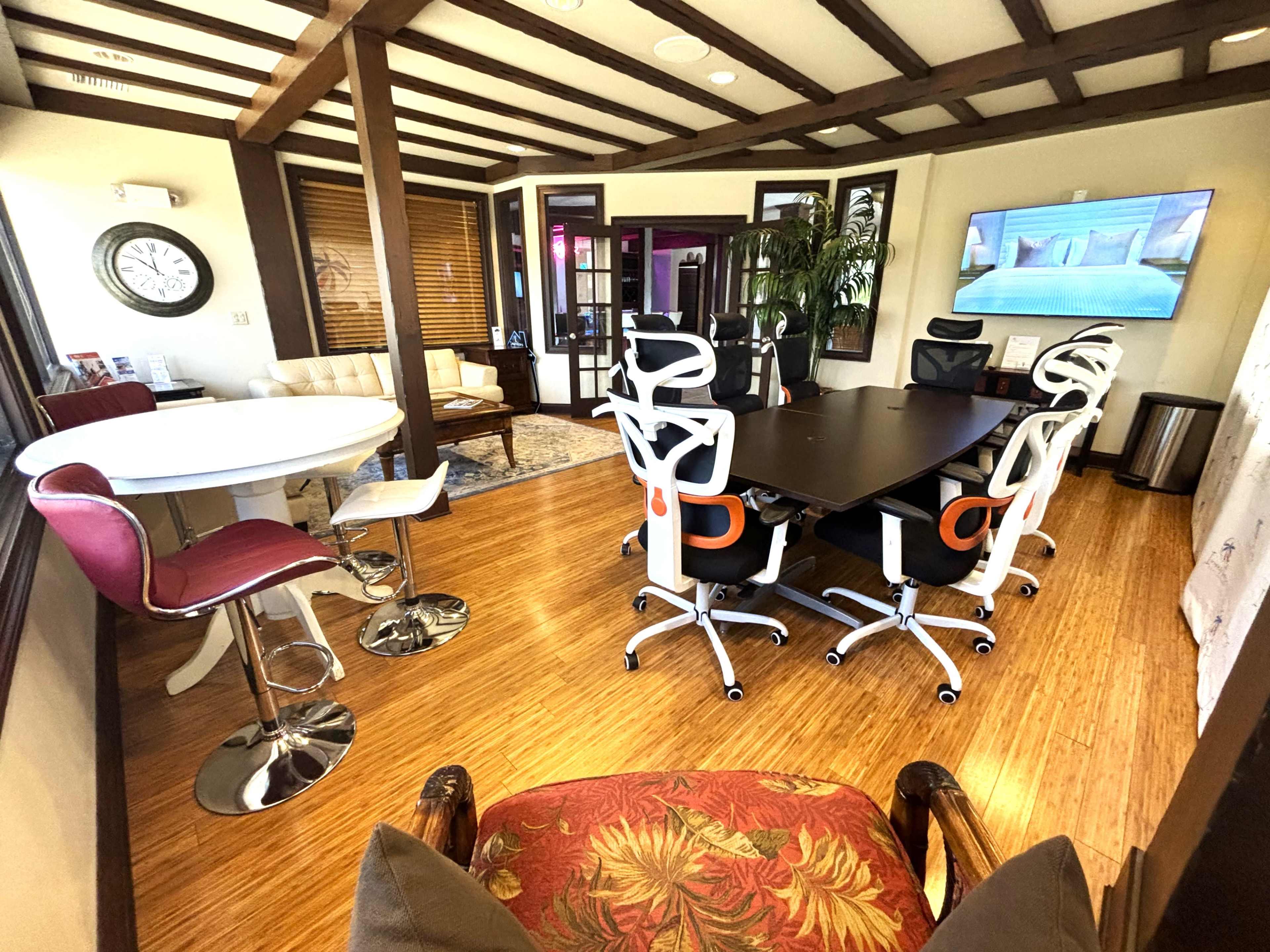 A modern conference room with a large black table surrounded by white ergonomic chairs, a seating area with a white sofa, and a flat-screen TV mounted on the wall.