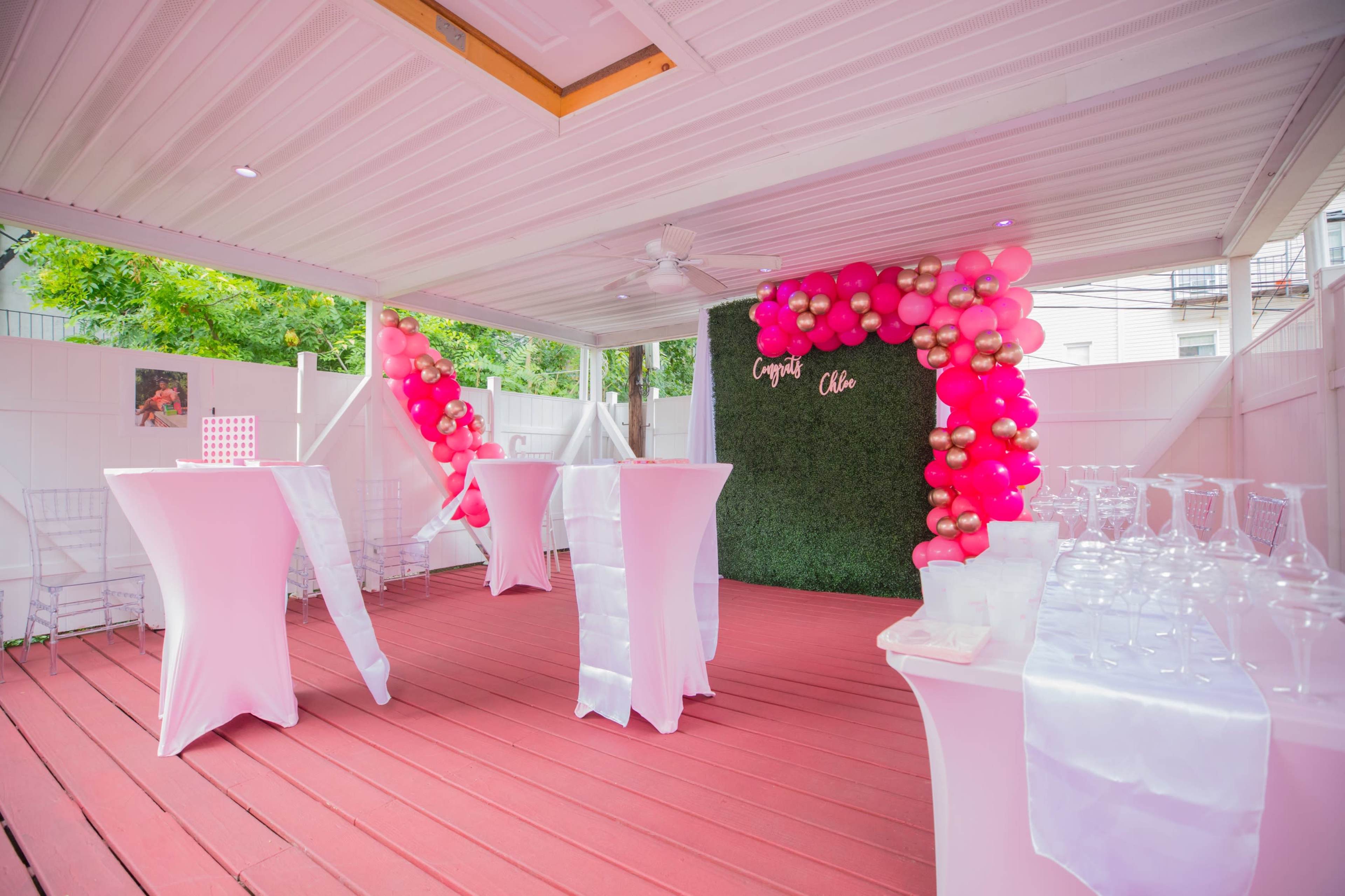 A decorated outdoor event space featuring pink and gold balloon arrangements, white cocktail tables, and a greenery backdrop.