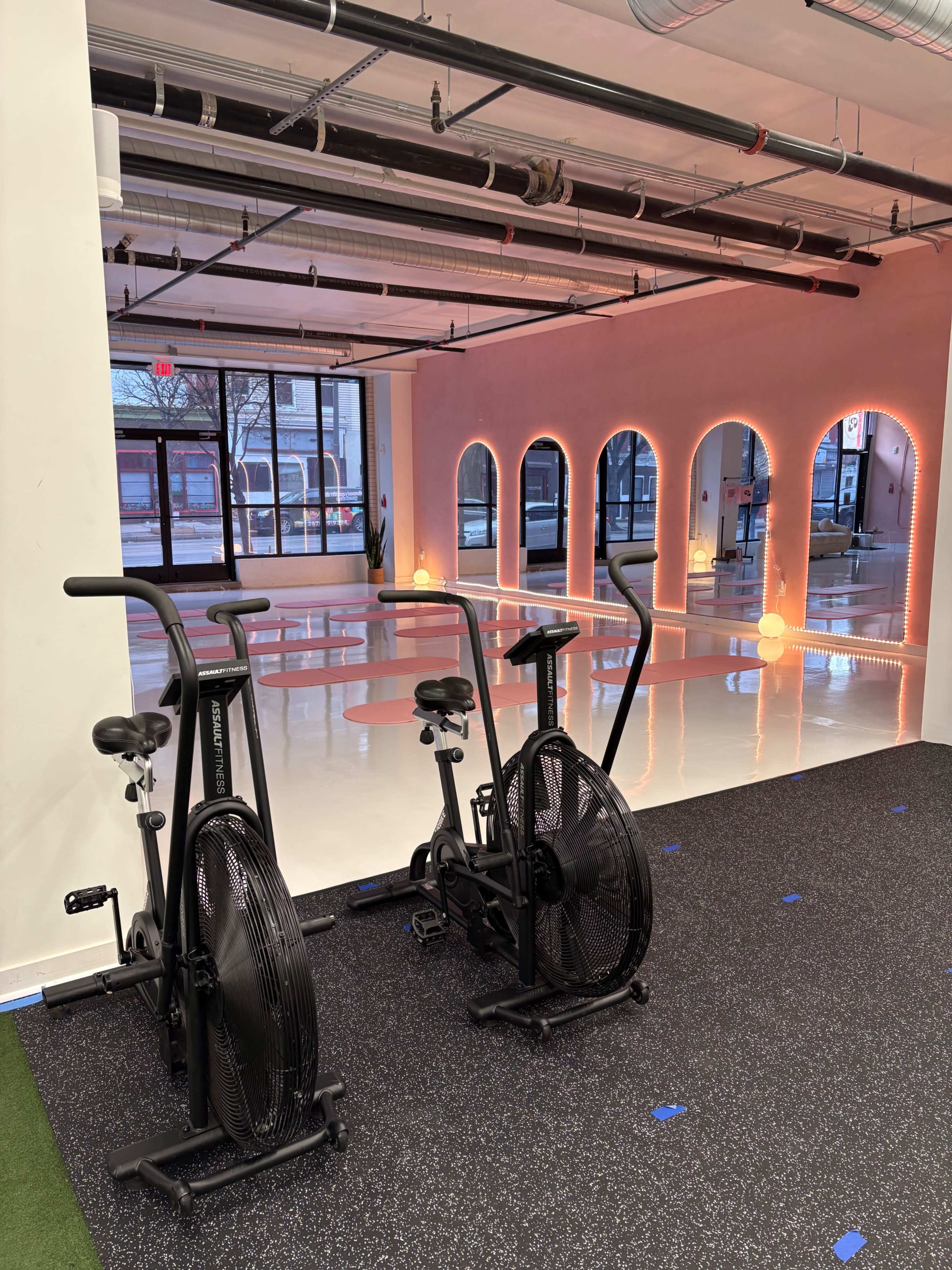 The image shows a fitness area with two stationary bikes in the foreground and a brightly lit, polished floor space featuring arches in the background.