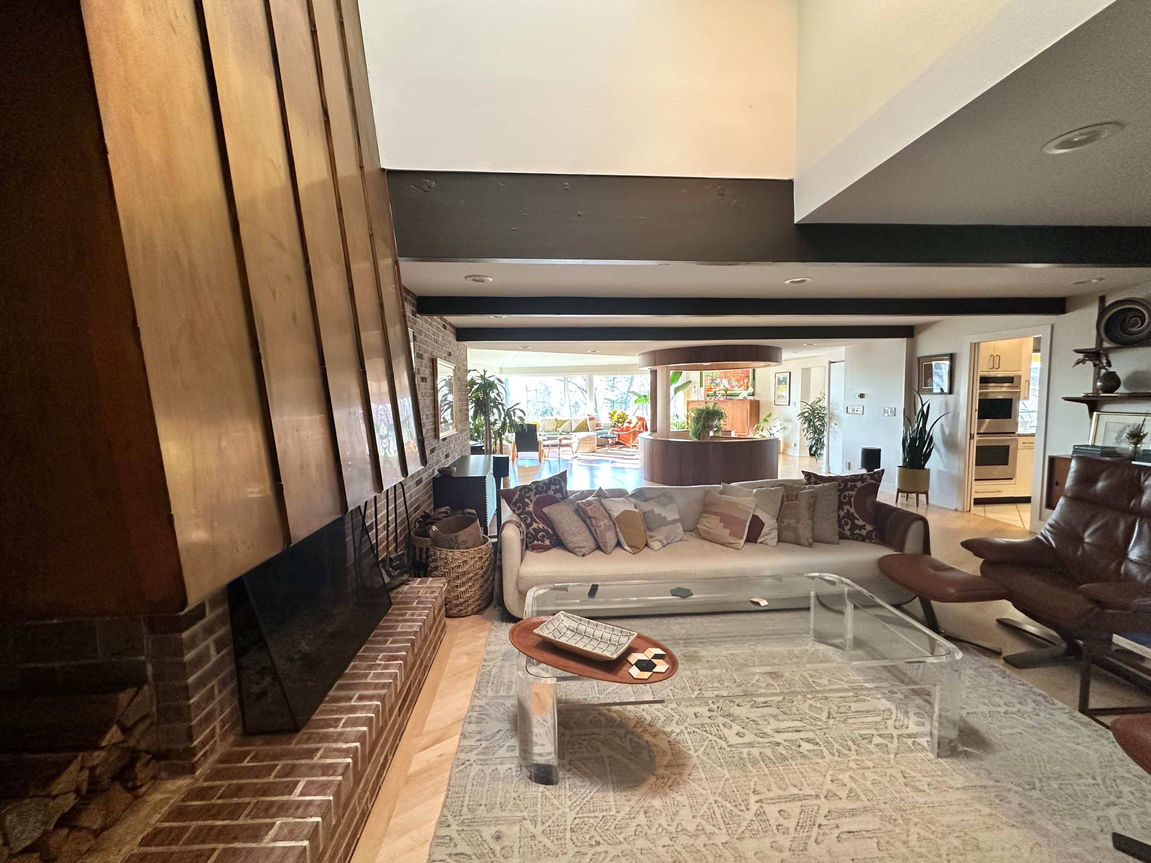 A living room with a sectional sofa, a glass coffee table, and a large brick fireplace, overlooking an open space with plants and kitchen area in the background.