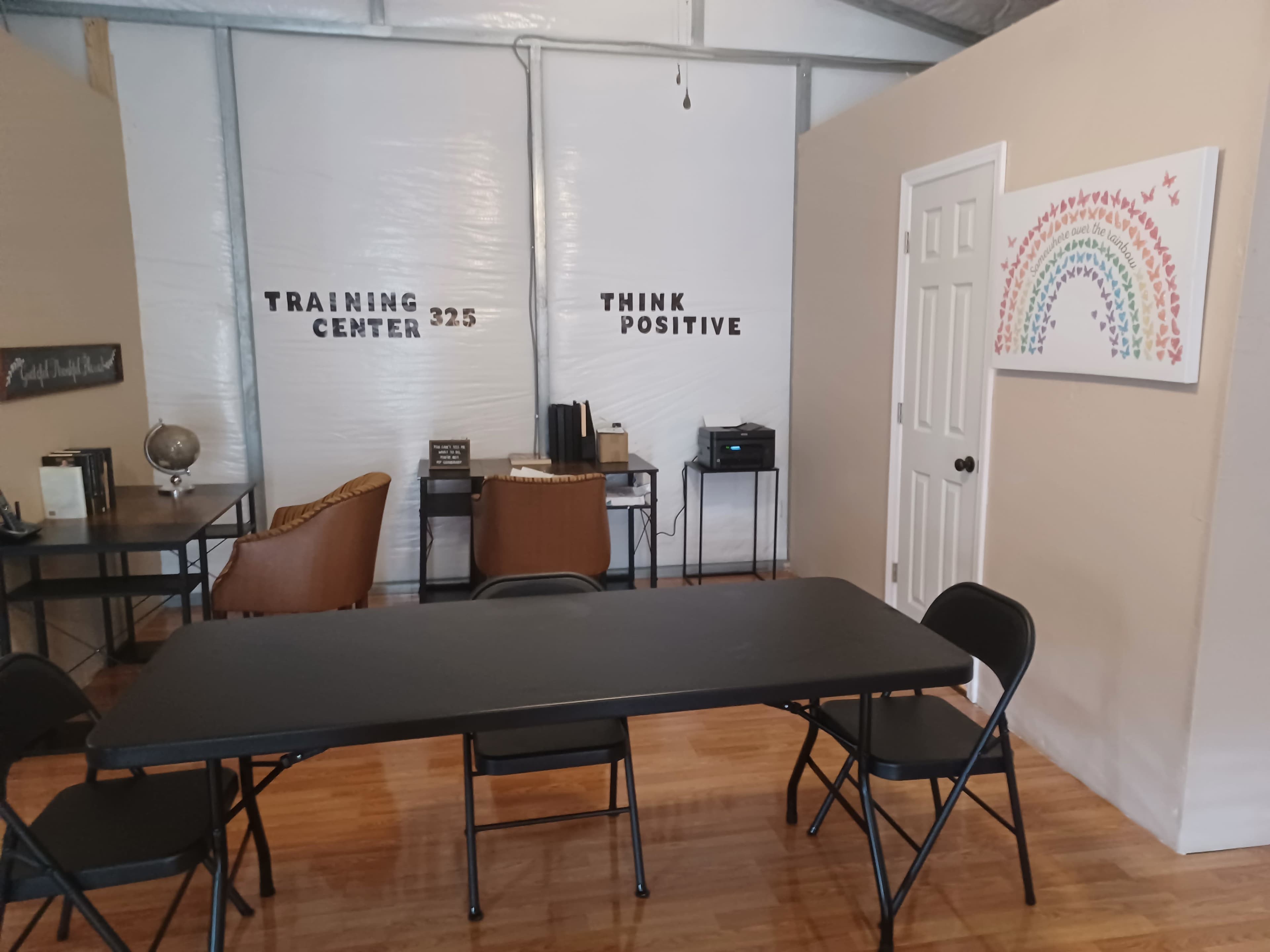 The image shows a room labeled "Training Center 325" with a table and chairs, a wall-mounted artwork depicting a rainbow, and an inspirational message that reads "Think Positive."